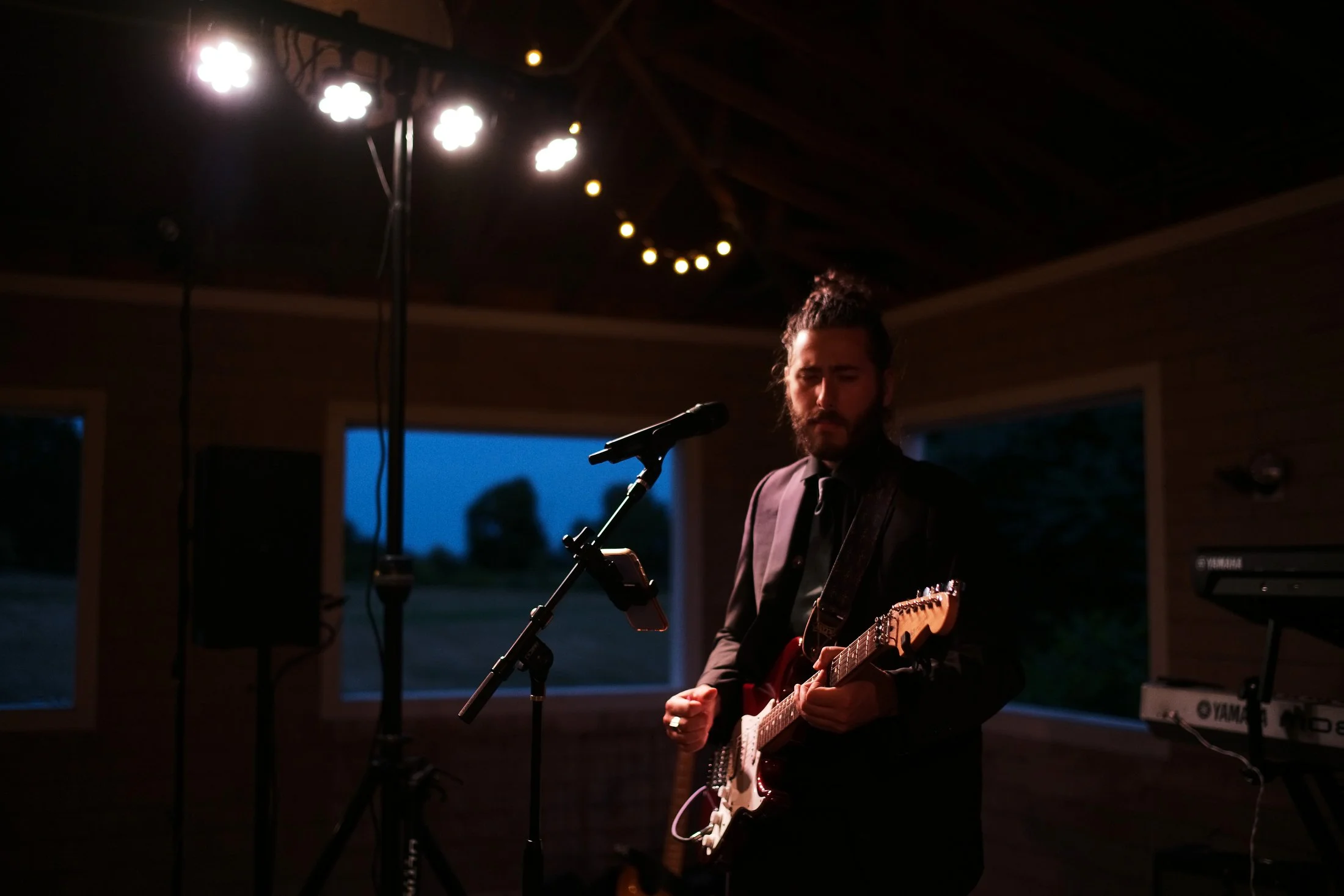 A man with a beard and long hair wearing a suit playing an electric guitar while standing in a dimly lit indoor space with large windows showing an evening sky outside.