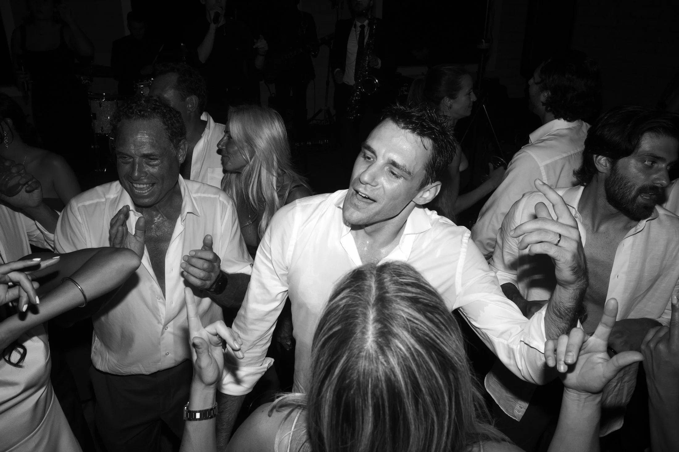 People dancing and celebrating at a party, with three men in the foreground smiling and engaging with the crowd.