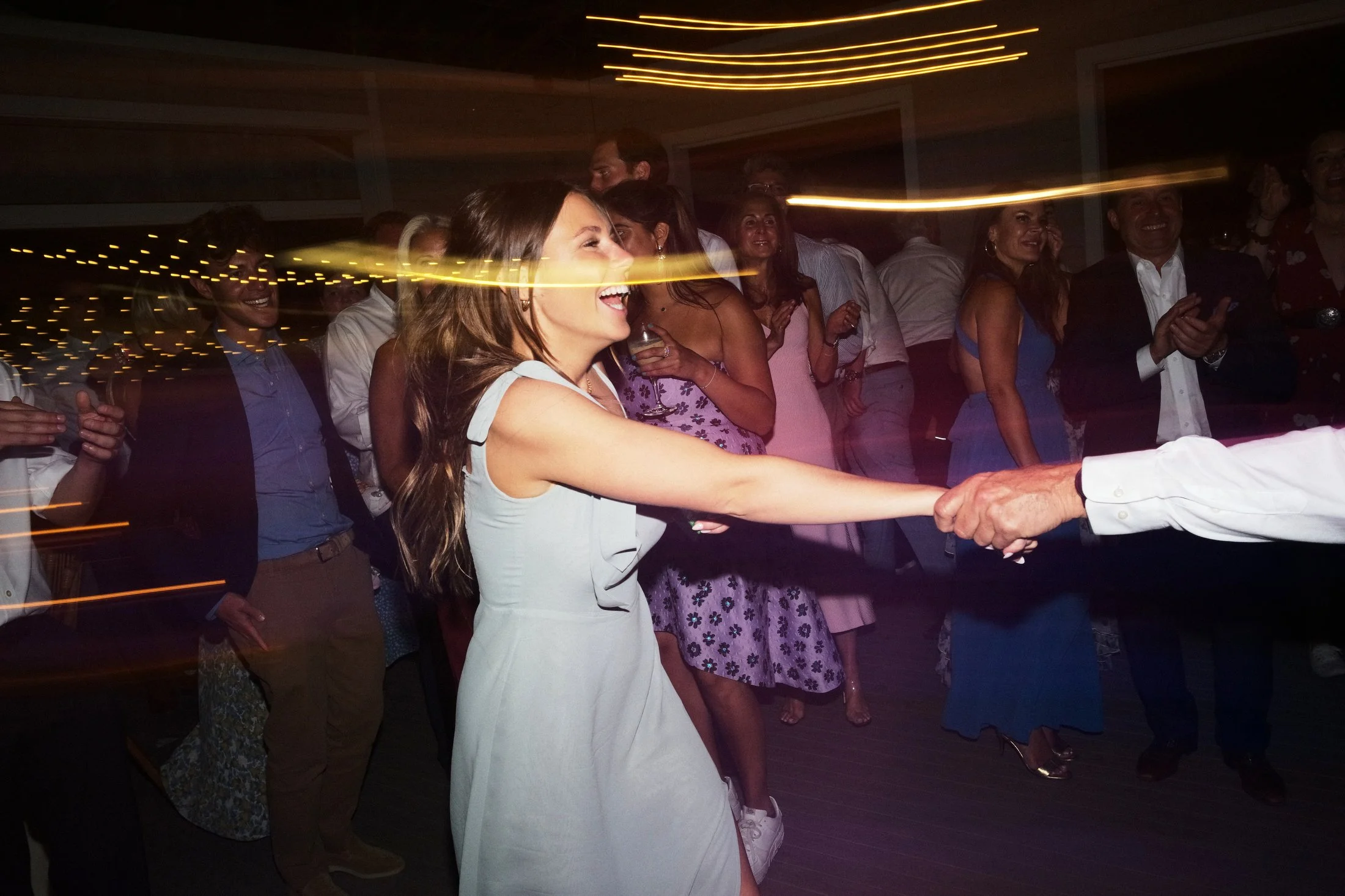 A woman in a white dress smiling and holding hands with a man during a dance at a party or celebration with other people watching and enjoying the moment.