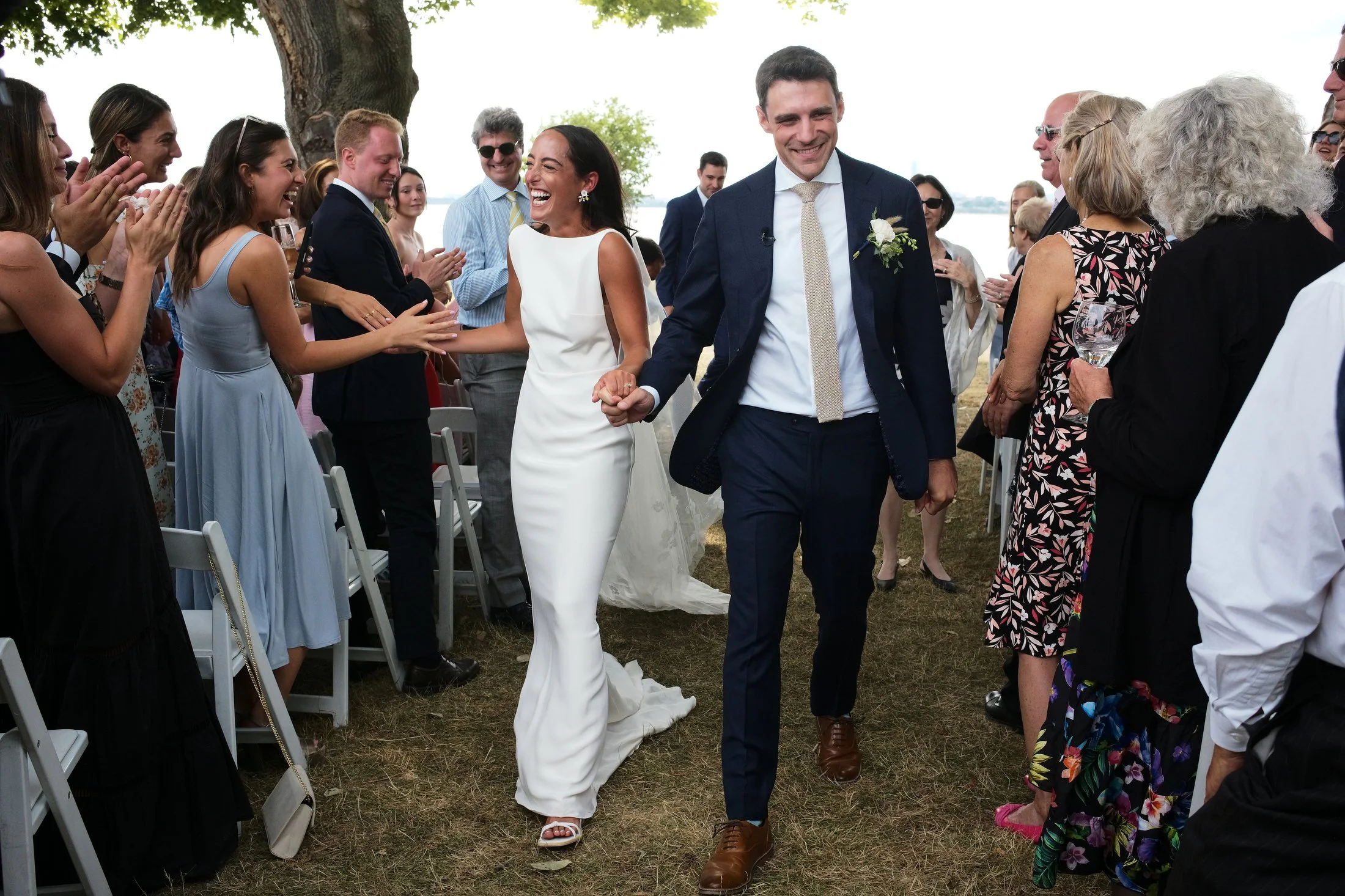 Bride and groom holding hands and smiling as they walk through a crowd of wedding guests outdoors by a tree, with water in the background.