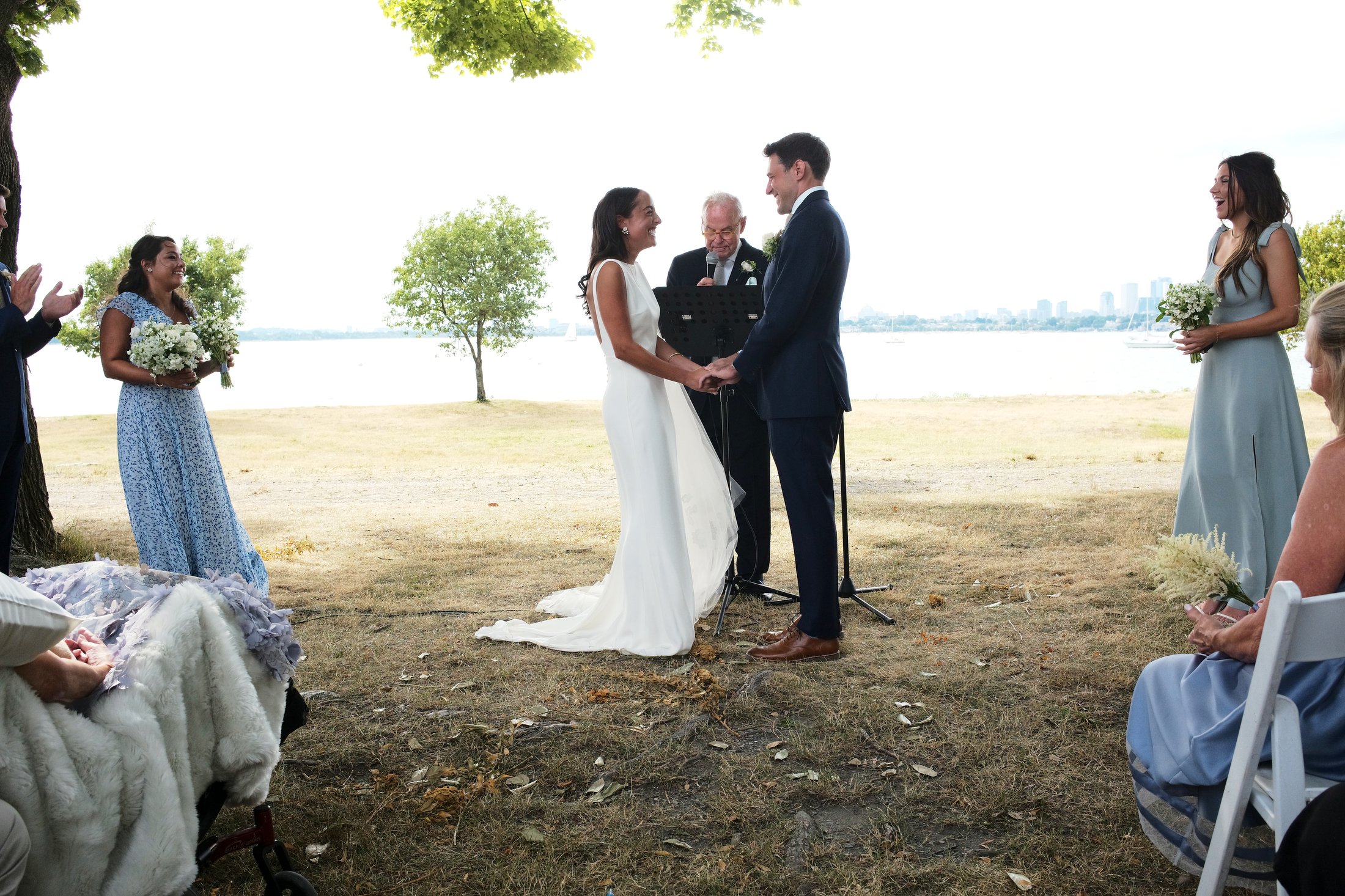 A couple gets married outdoors by a body of water, holding hands and facing each other, with an officiant reading from a booklet. Bridesmaids and guests surround them, with trees and a city skyline in the background.