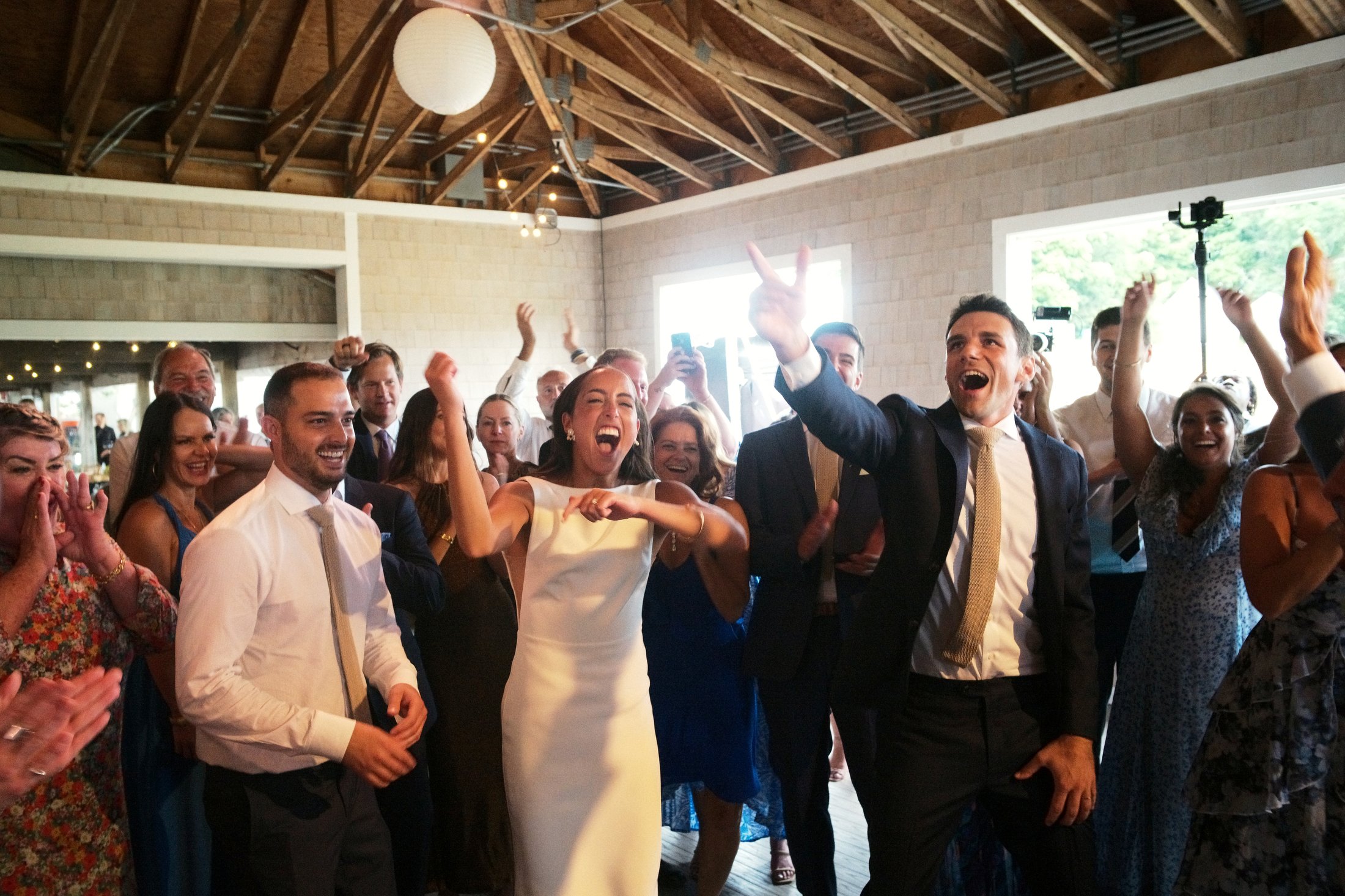 Group of people at a celebration, seen dancing and smiling indoors with high wooden ceilings and large windows, dressed in formal attire.