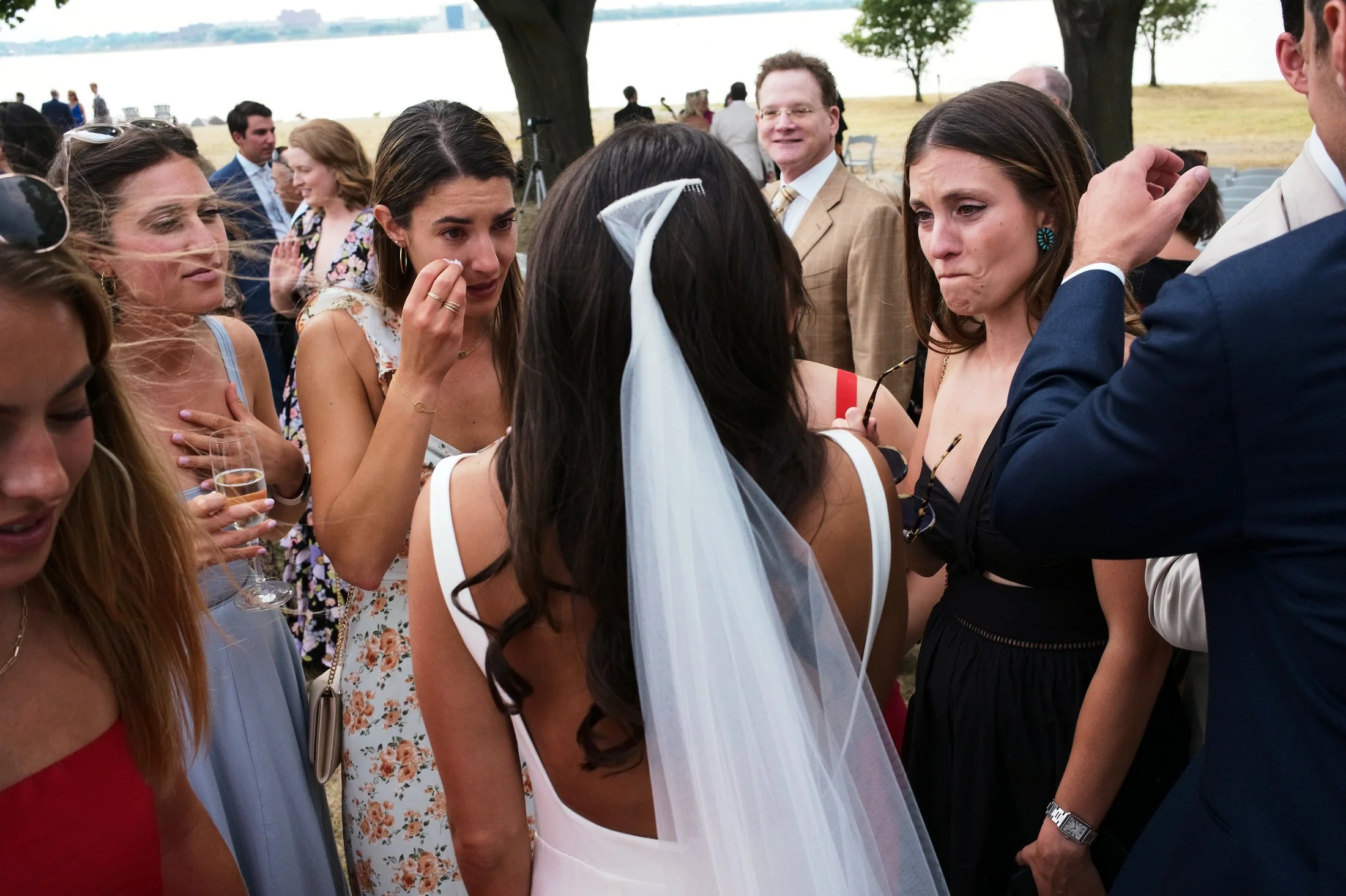 A woman in a white wedding dress and veil is surrounded by a group of women and men at an outdoor wedding reception. The women appear emotional, some crying, and one is wiping her tears. The setting is near a body of water with trees and grassy area 