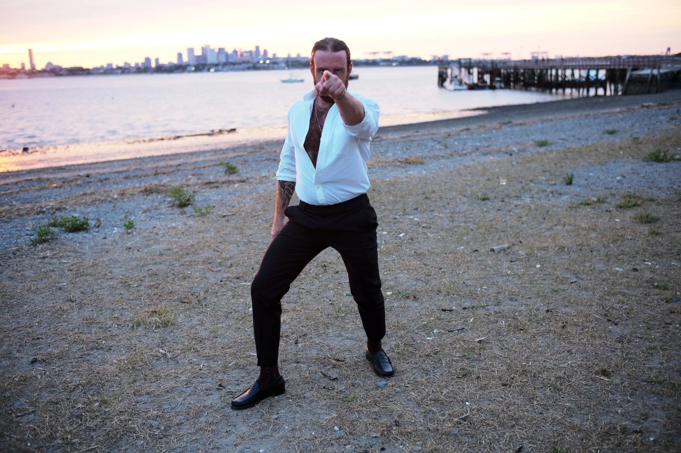 A man with dark hair and tattoos on his arm, wearing a partially unbuttoned white shirt and black pants, stands on a beach at sunset, pointing directly at the camera.