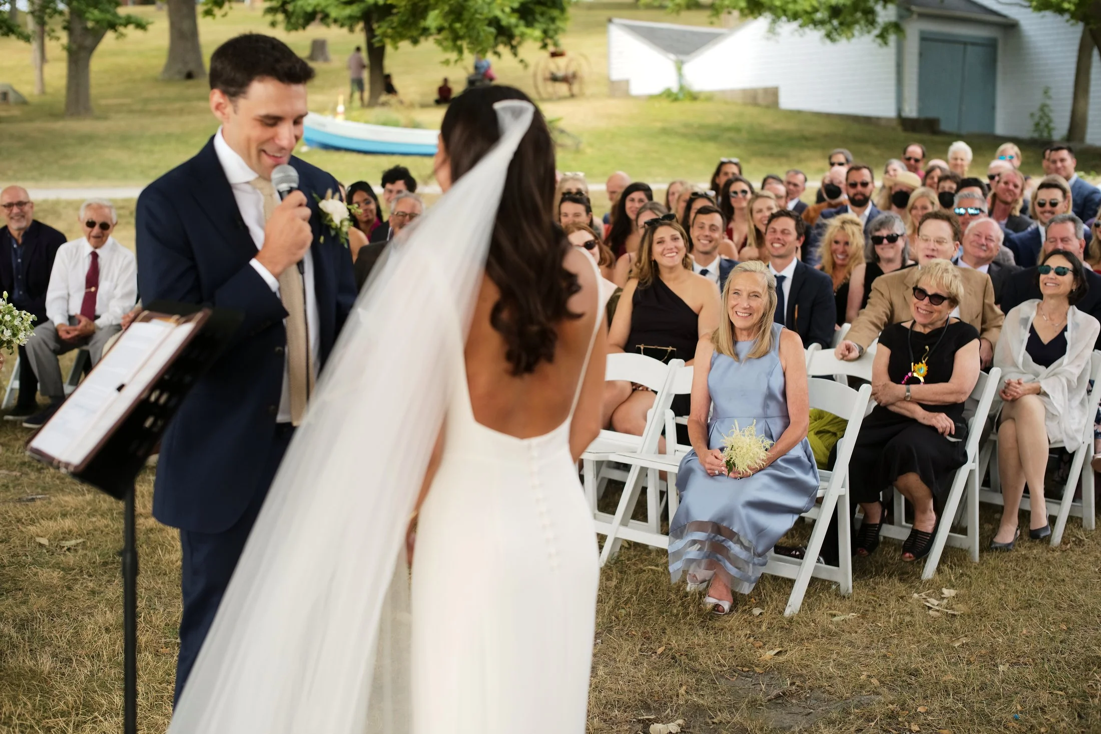A bride and groom exchanging vows in an outdoor wedding ceremony, with guests seated on white chairs in the background, smiling and watching the event.
