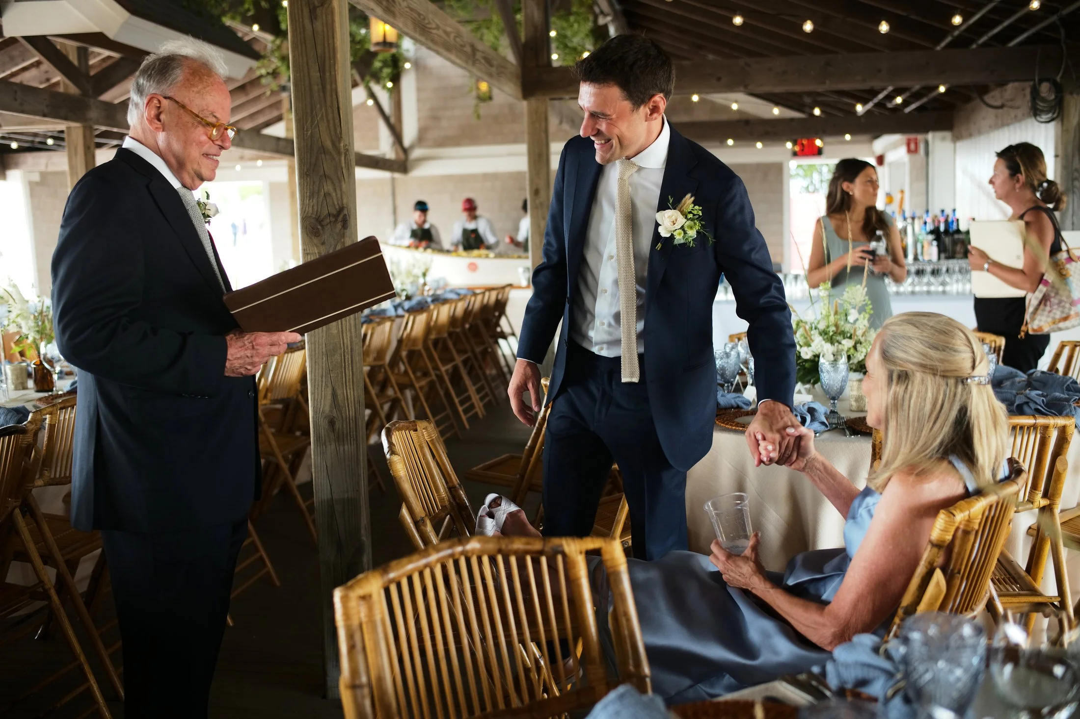 A wedding reception with a groom holding the hand of a seated bride, looking at an older man reading a card, with women in the background.