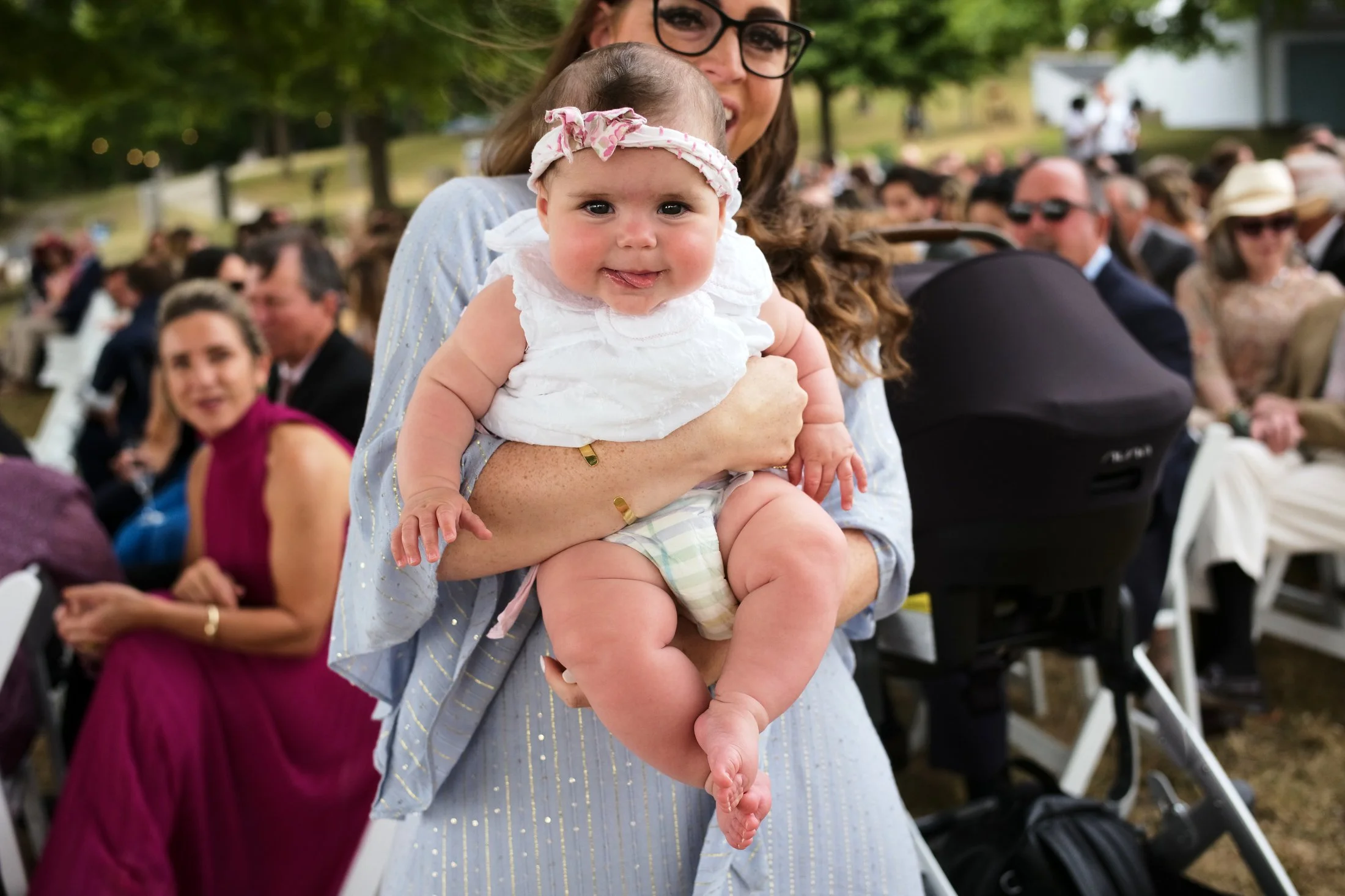 A woman holding a smiling baby girl at an outdoor event with many people seated in the background.
