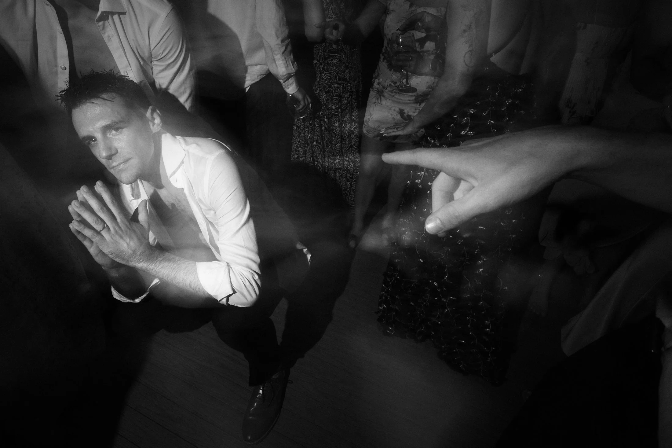 A man in a white shirt with rolled-up sleeves is kneeling with hands pressed together in a prayer-like gesture at a party. There are other people around him, some holding drinks, and the scene is dimly lit with a black-and-white filter.