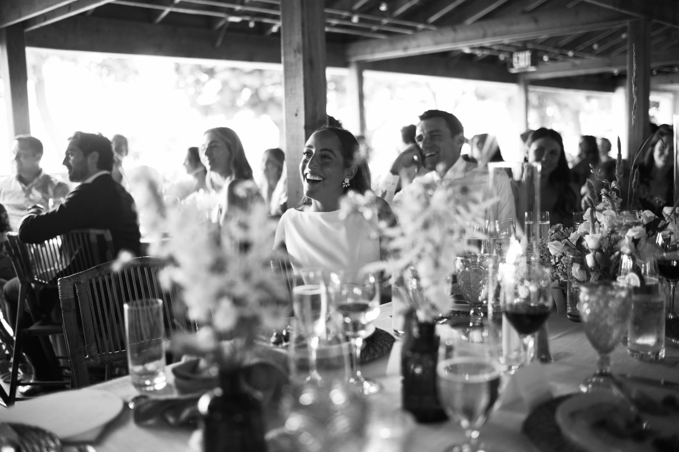 Guests at a wedding reception sitting at a table, smiling and laughing, with floral centerpieces and drinks in glasses, in a rustic outdoor setting with a wooden structure.