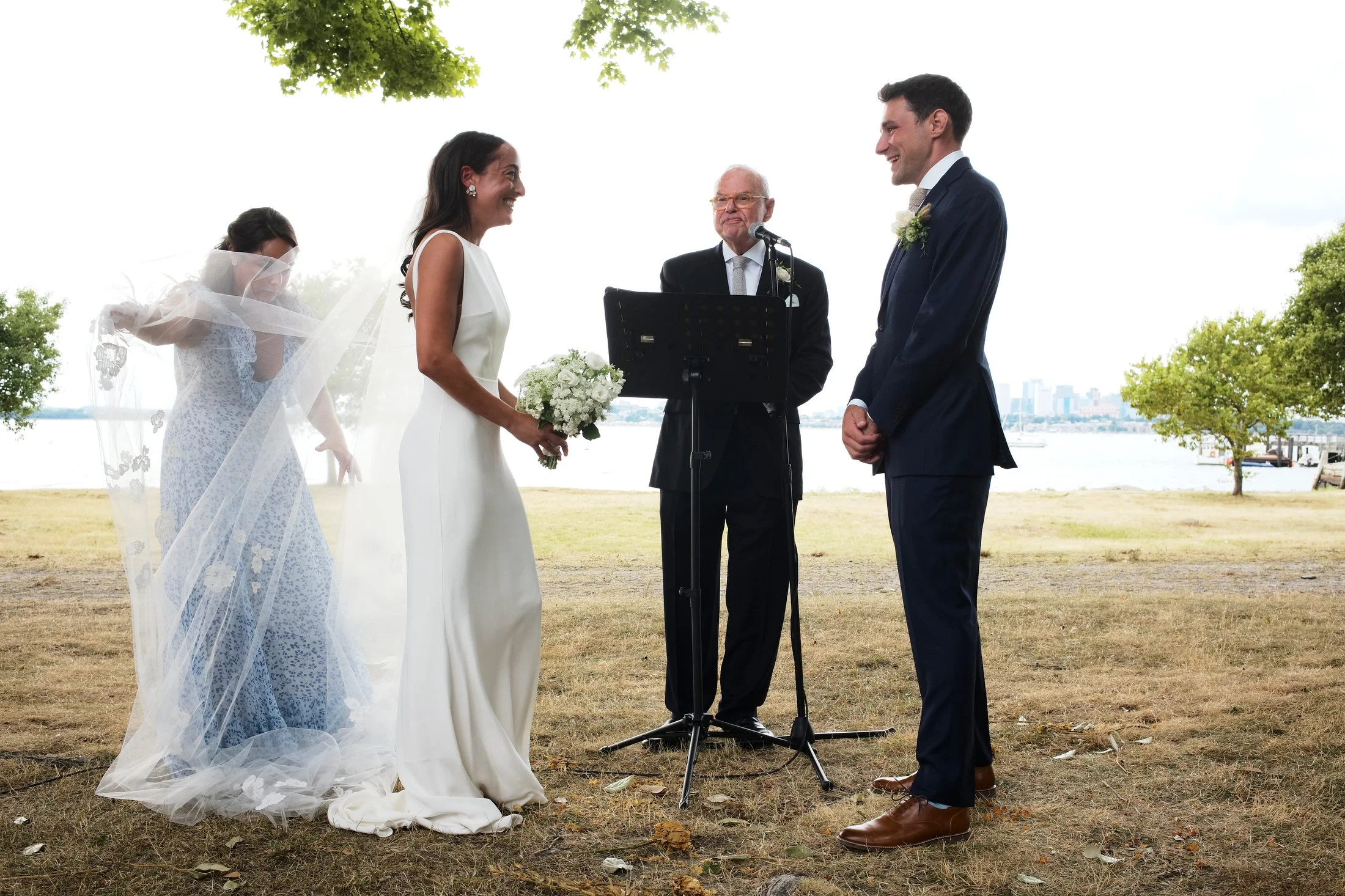A wedding ceremony outdoors near a body of water with trees and city skyline in the background. A bride and groom are facing each other, exchanging vows, while an officiant stands between them. The bride wears a white dress and holds a bouquet, and t