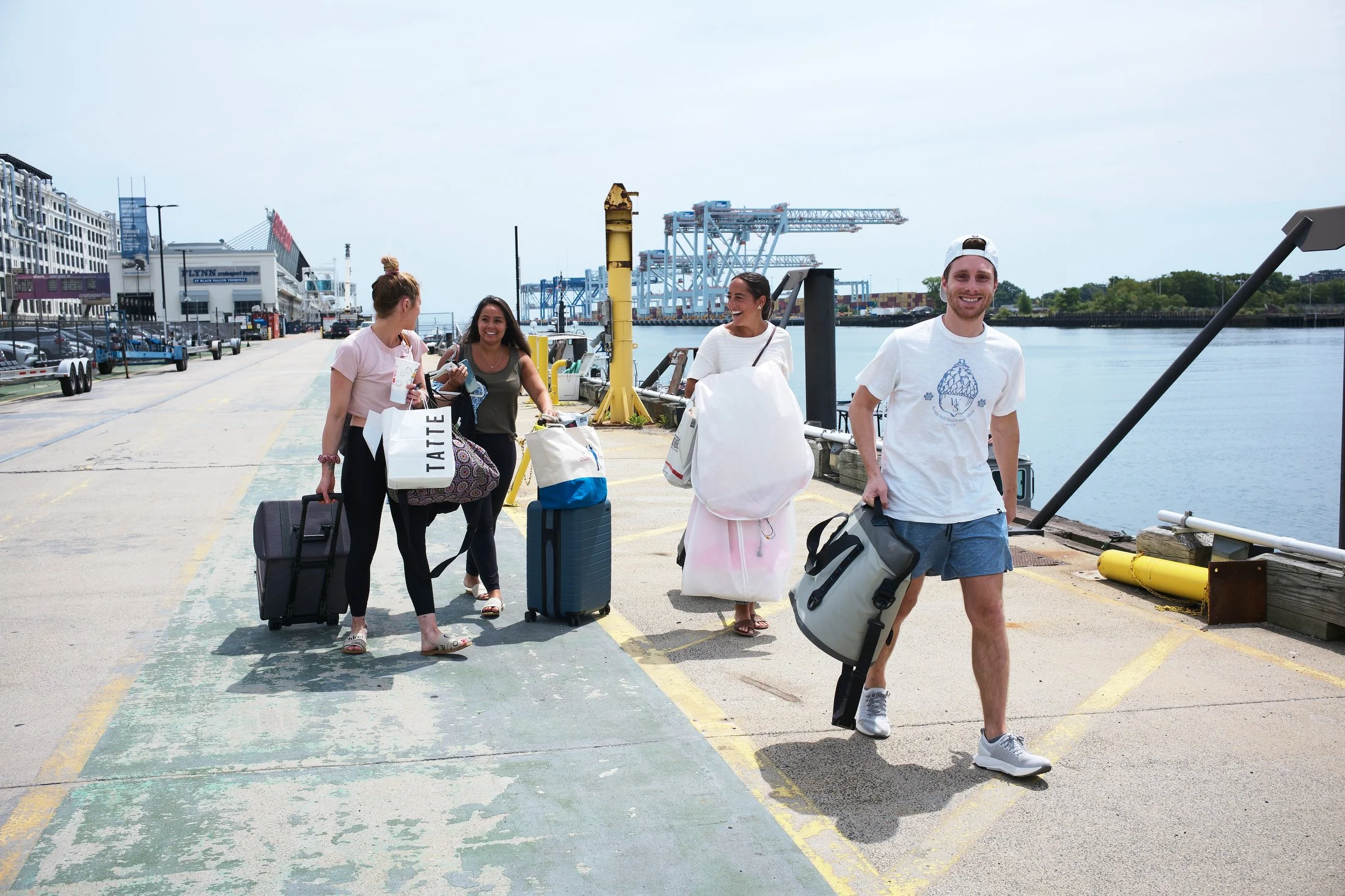 Group of four young adults, two women and two men, walking along a dock with luggage and bags, near water and industrial cranes in the background, on a partly cloudy day.