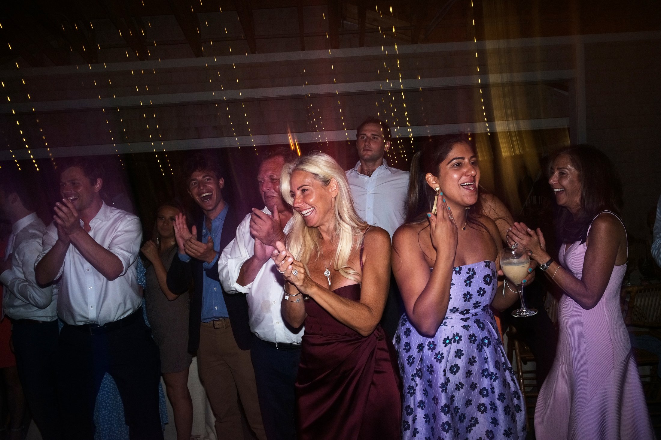 A group of people enjoying a celebration, dancing and clapping, at an indoor party with string lights overhead. Some women are dressed in colorful dresses, one woman is holding a drink, and everyone looks happy and engaged.