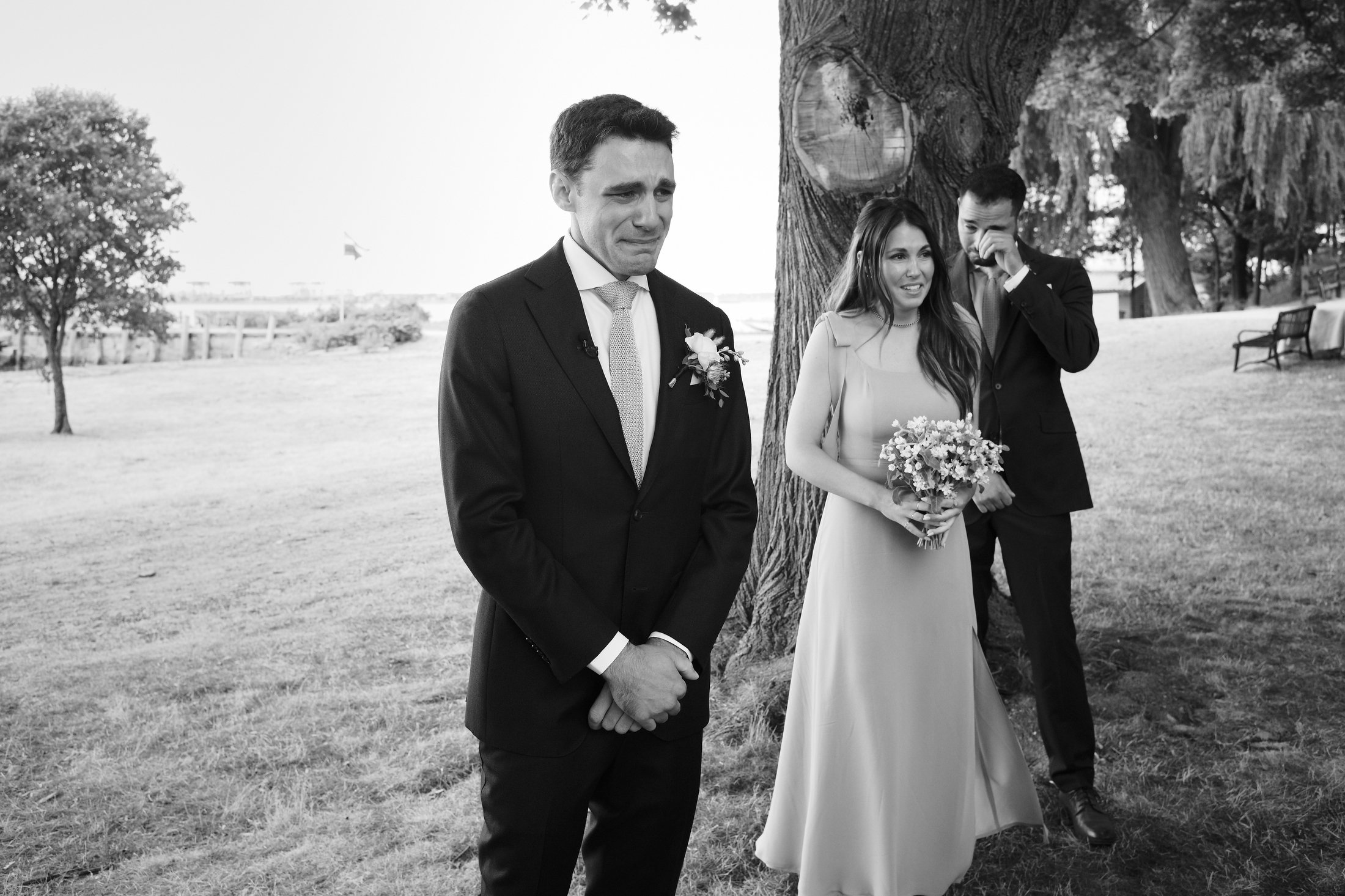 Black and white photograph of a wedding scene outdoors, with a bride holding a bouquet standing under a large tree and a groom crying, while a man wipes away tears.