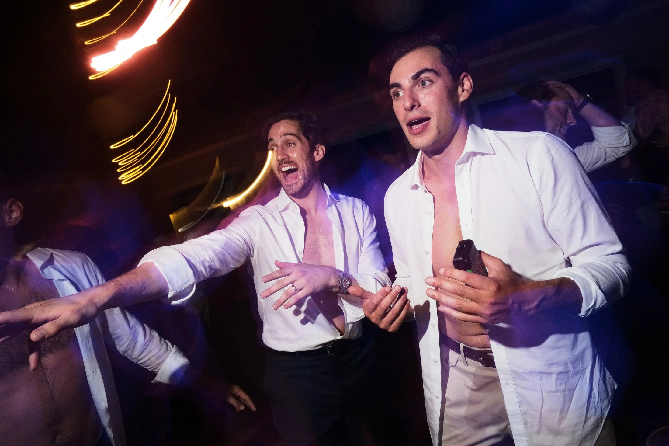 Two men in white shirts with their shirts unbuttoned at the top are partying together with others in a dark, colorful nightclub, with light streaks and blurred motion indicating dancing.