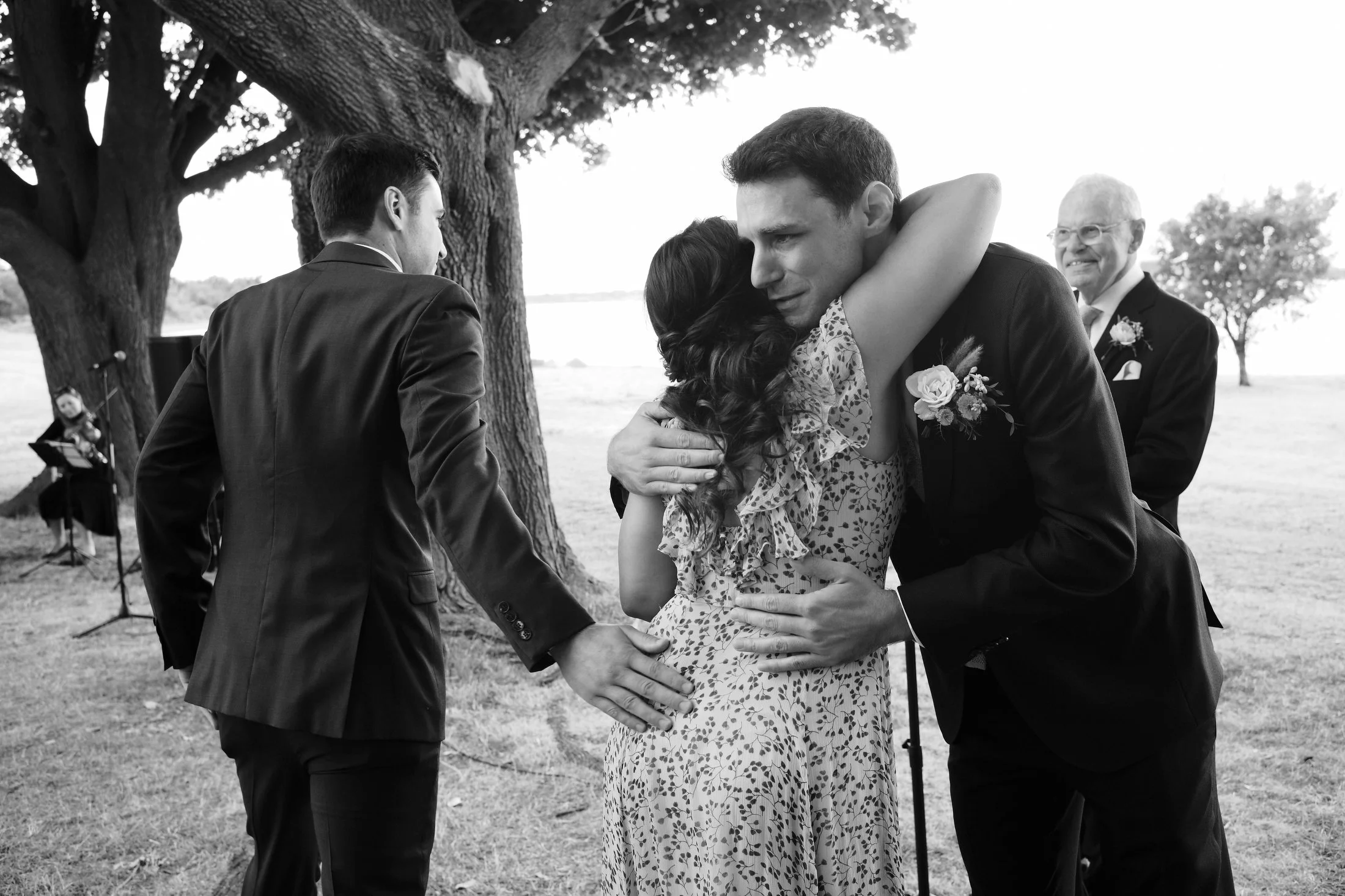 A black and white photo of a couple hugging during a wedding ceremony outdoors under a large tree, with another man standing nearby and an elderly man in the background.