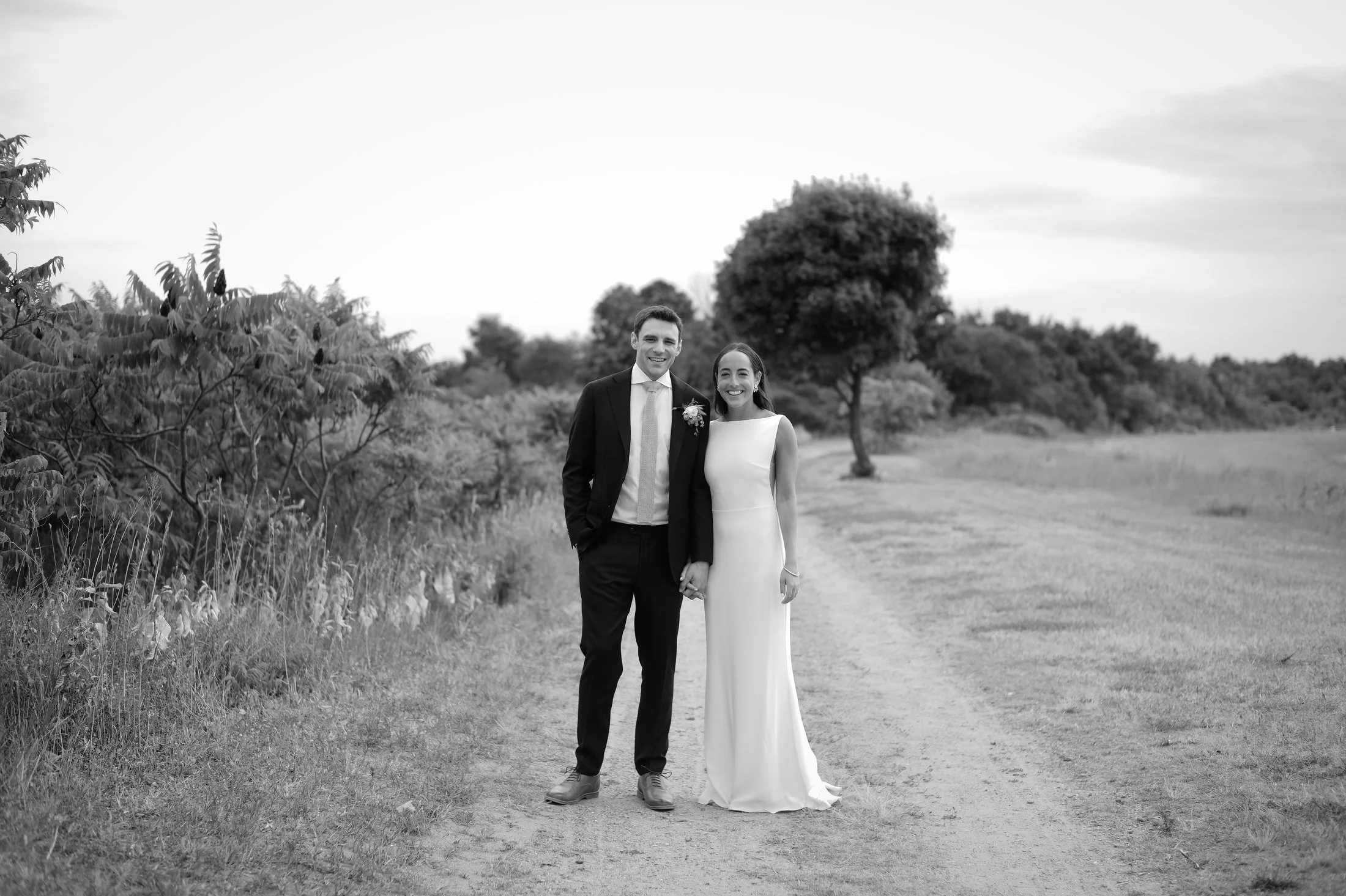 A black-and-white photo of a wedding couple standing hand-in-hand on a dirt path in a rural area with trees and grasses in the background, smiling at the camera.