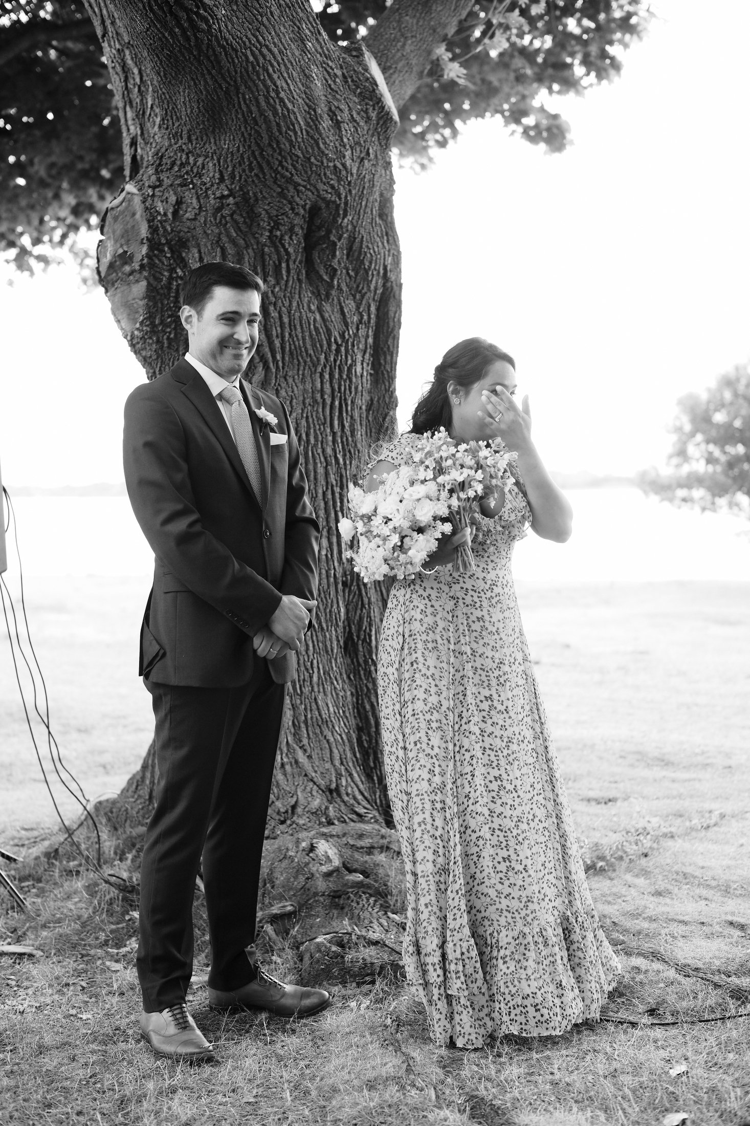 A black and white photo of a bride and groom standing outdoors near a large tree. The groom is smiling and dressed in a suit. The bride is holding a bouquet of flowers, covering her face with her hand, possibly wiping away tears.