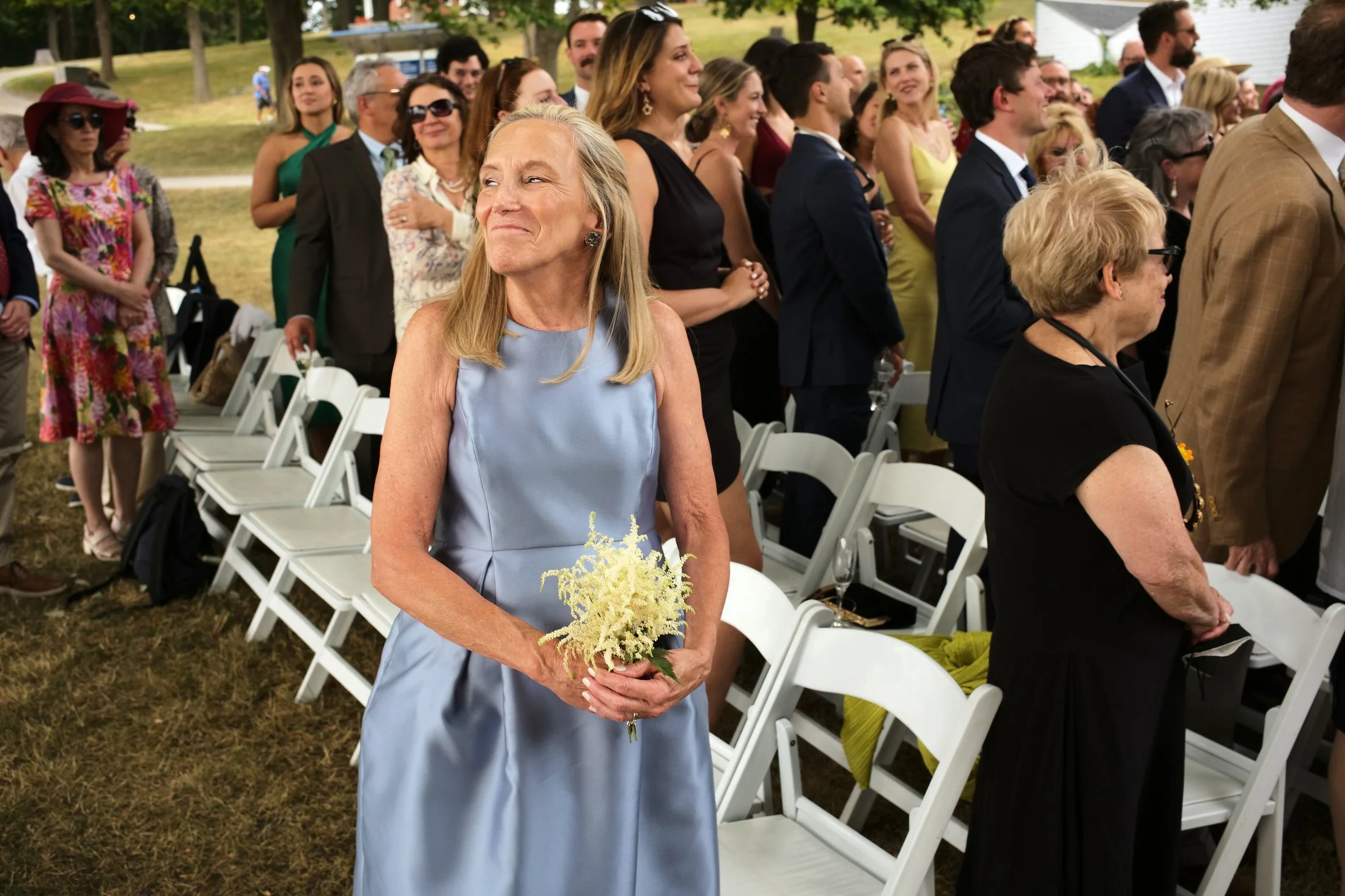A woman in a light blue dress holding a small bouquet of white flowers at a wedding ceremony outdoors, surrounded by seated and standing guests in formal attire.