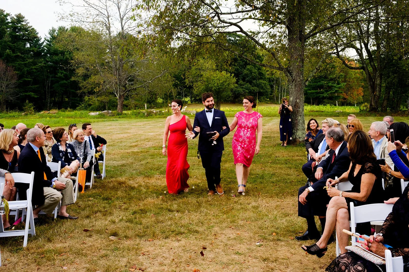 A wedding ceremony outdoors with guests seated on either side of a grassy aisle. A man in a tuxedo and two women in pink dresses walk down the aisle, smiling. Trees and green landscape are in the background.