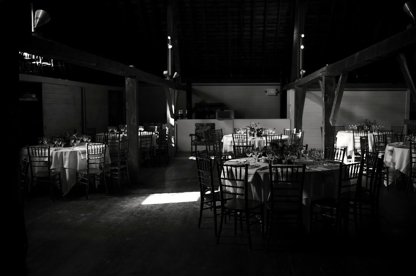Empty banquet hall with round tables covered with white tablecloths, set with glassware and silverware, illuminated by sunlight through windows.