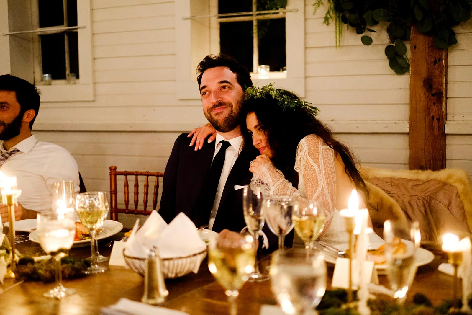 A couple at a wedding reception sitting at a table, with the woman resting her head on the man's shoulder, surrounded by candles and glasses of wine.