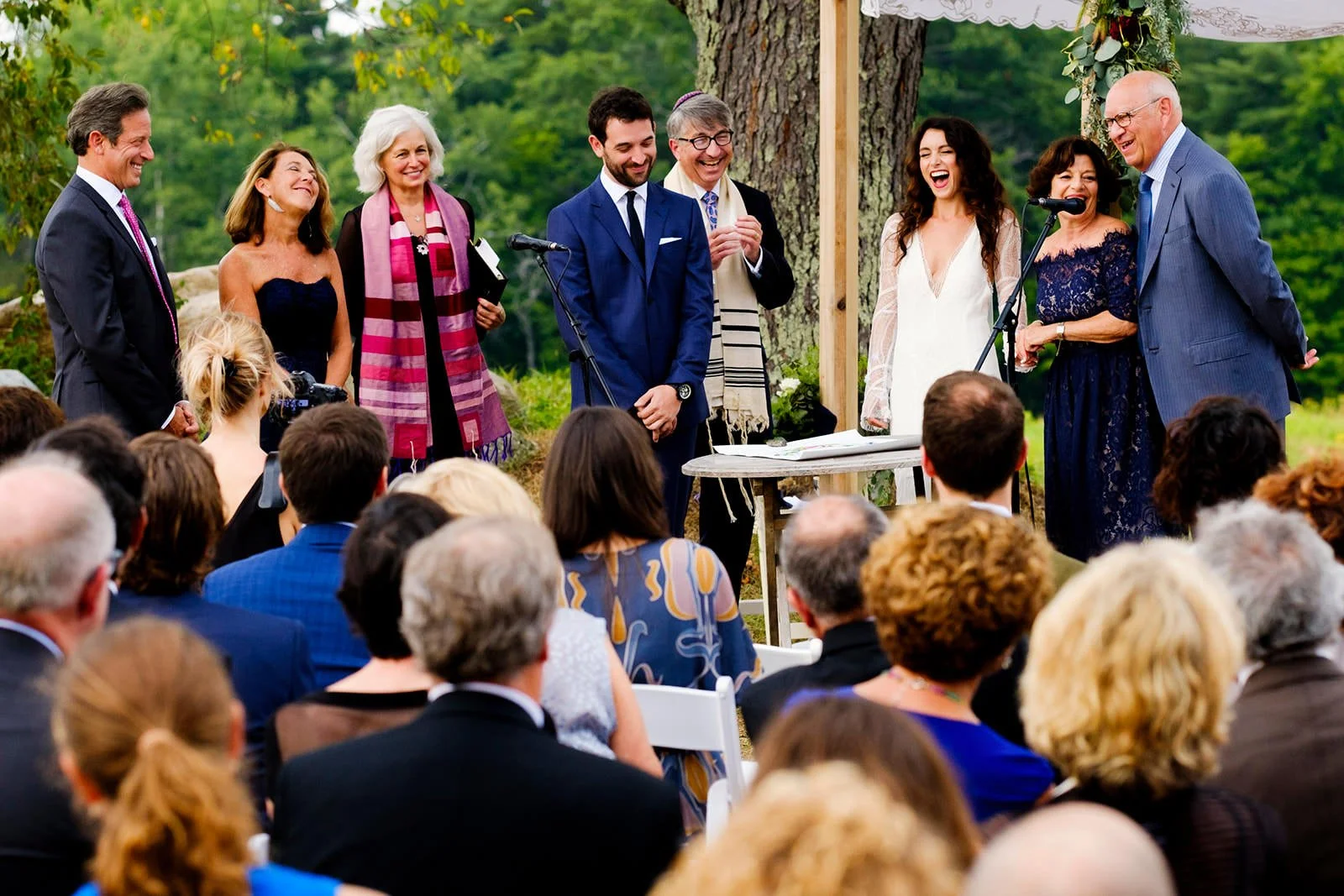 People at an outdoor wedding ceremony, smiling and laughing, with a large tree and green scenery in the background