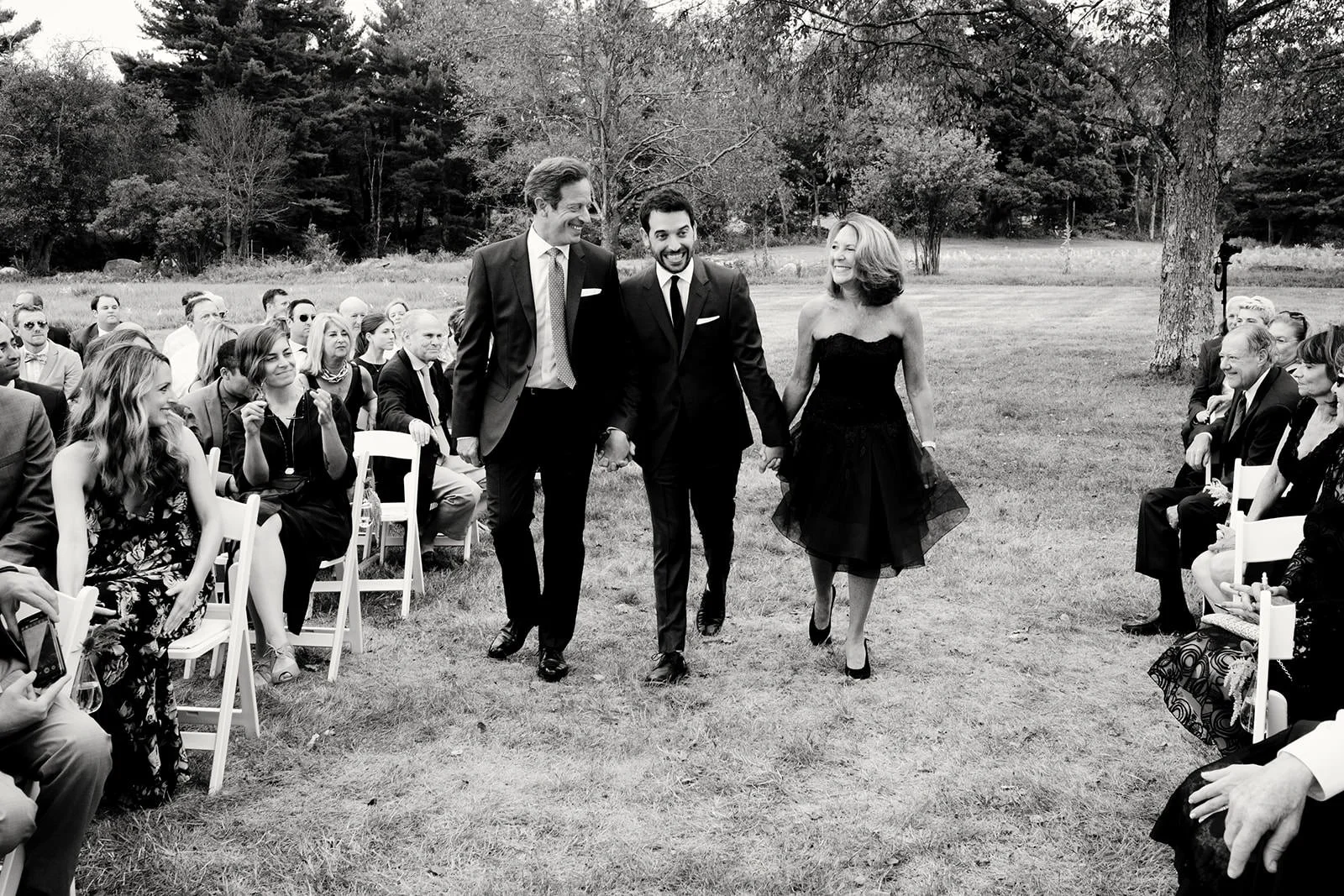 A black-and-white photo of a wedding ceremony outdoors, with a group of guests seated on chairs on either side. A couple, a man and a woman, are walking down the aisle holding hands, dressed in formal attire with the woman in a black dress and the me