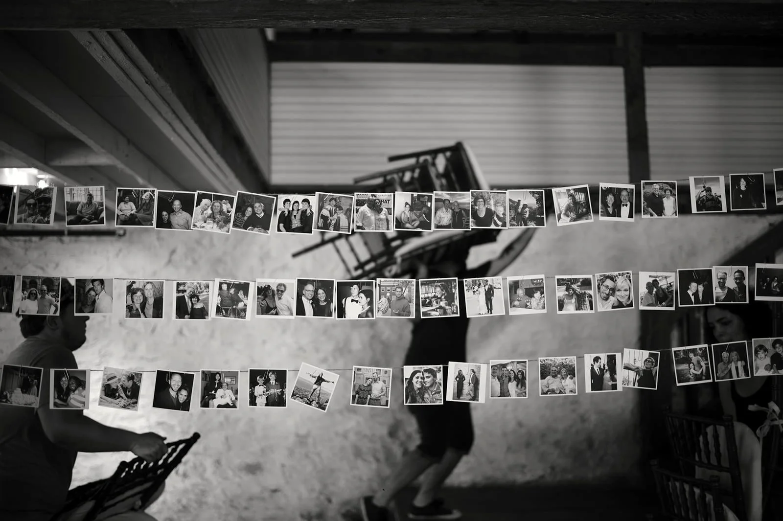 Black and white photo of string photo displays with various pictures hung in an indoor setting, with a person in the background, and part of a person sitting on the left side.