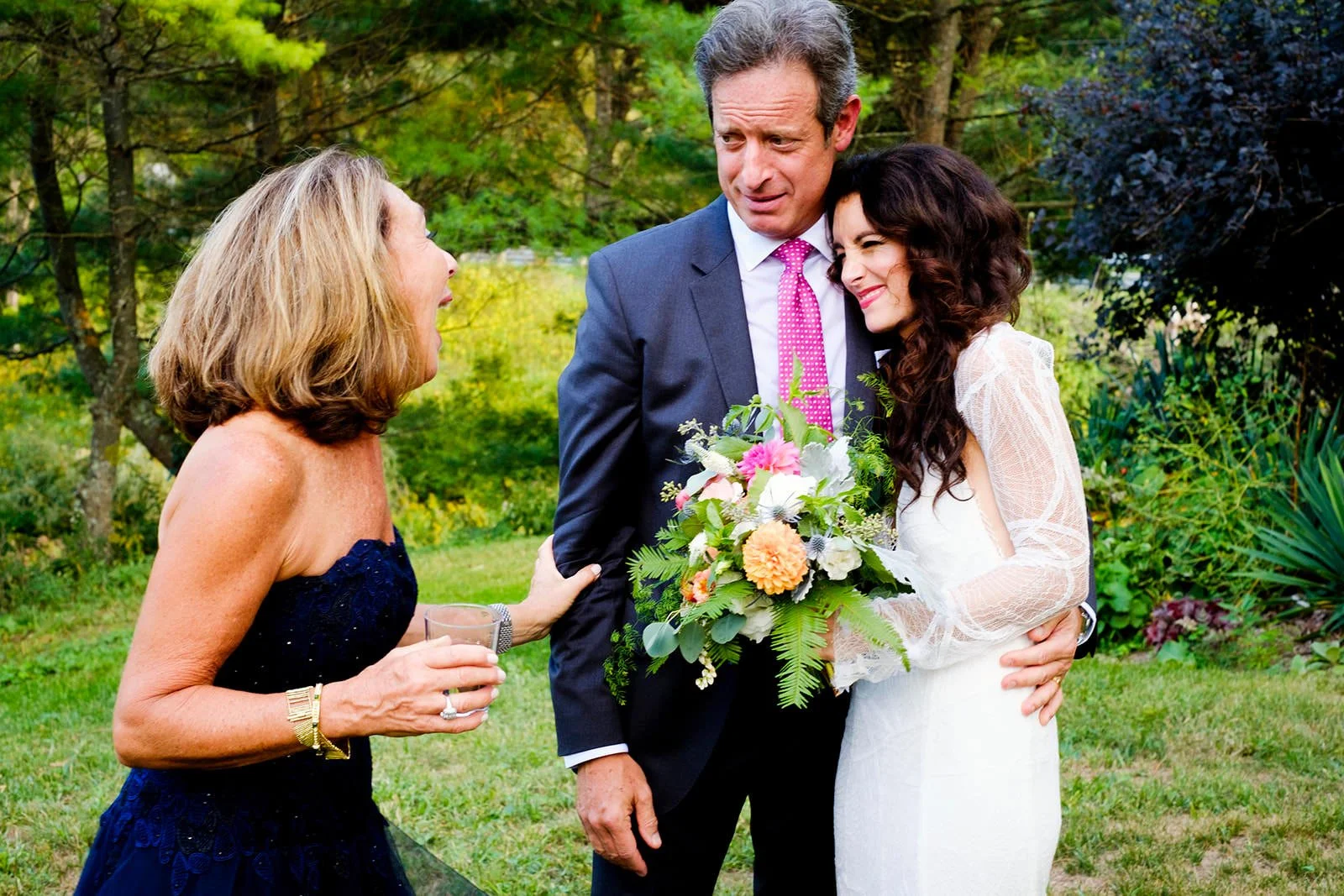A bride in a white wedding dress and a man in a navy suit with a pink tie are hugging and smiling, while a woman in a dark blue strapless dress holding a glass and gesturing emotionally talks to them outdoors in a garden.