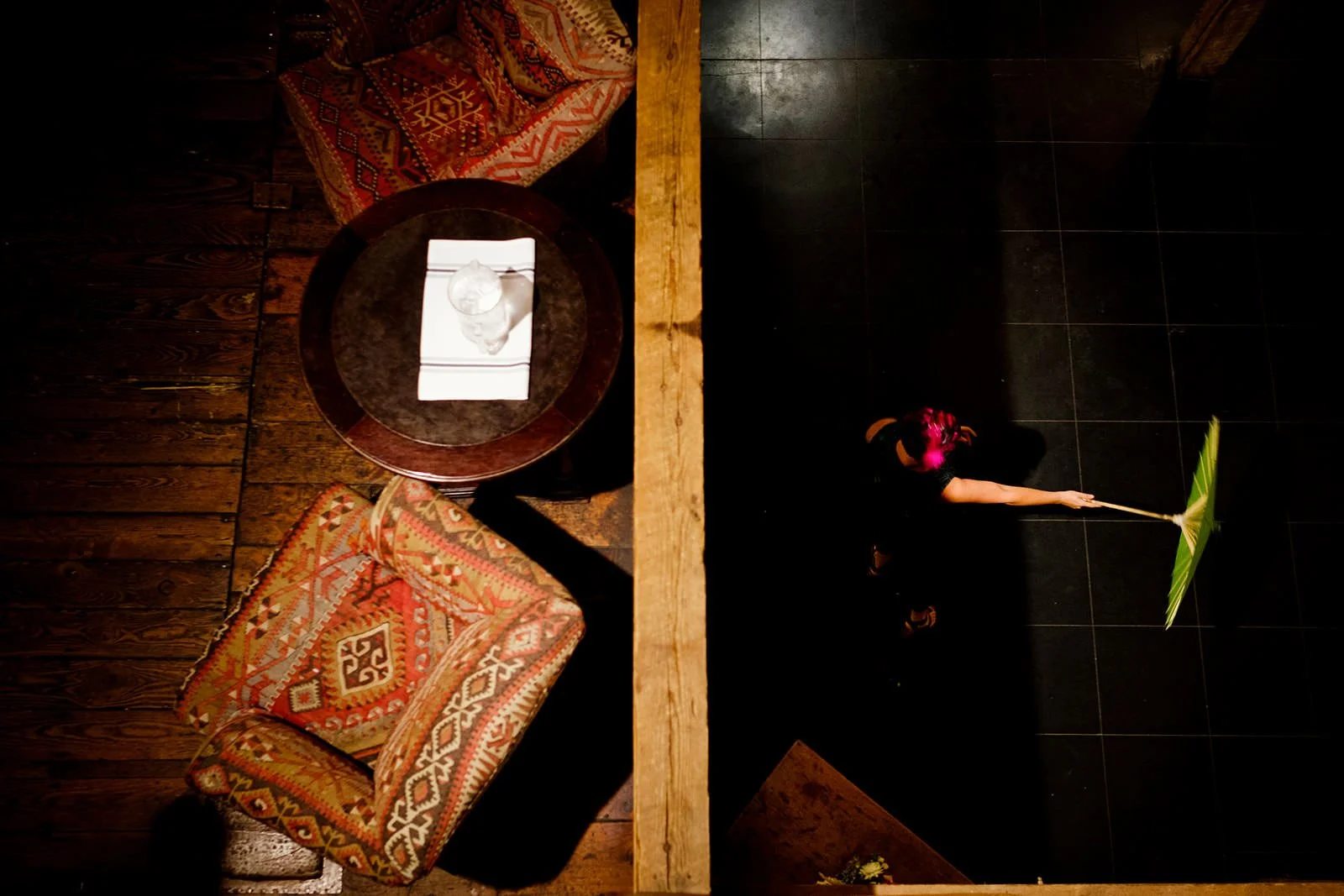 Top-down view of a cozy indoor space divided by a wooden beam. On the left side, there are two patterned armchairs with red, orange, and beige designs, along with a round wooden table holding a water bottle on top of a white cloth. The right side fea