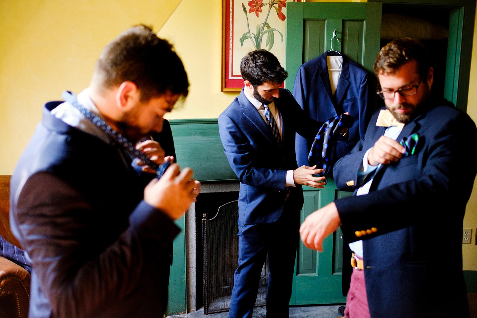 Group of men in suits adjusting their bow ties and ties in a room with yellow walls and a decorative painting.