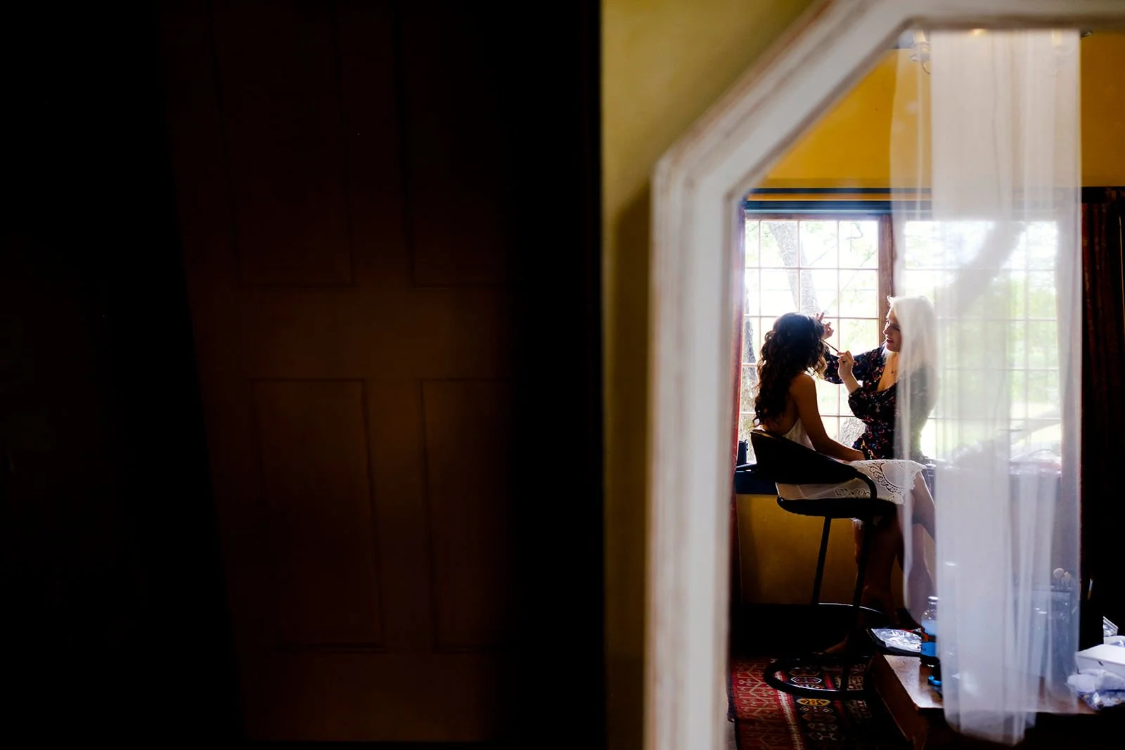 Two women are sitting by a large window, where one is applying makeup to the other in a bright, homey room.
