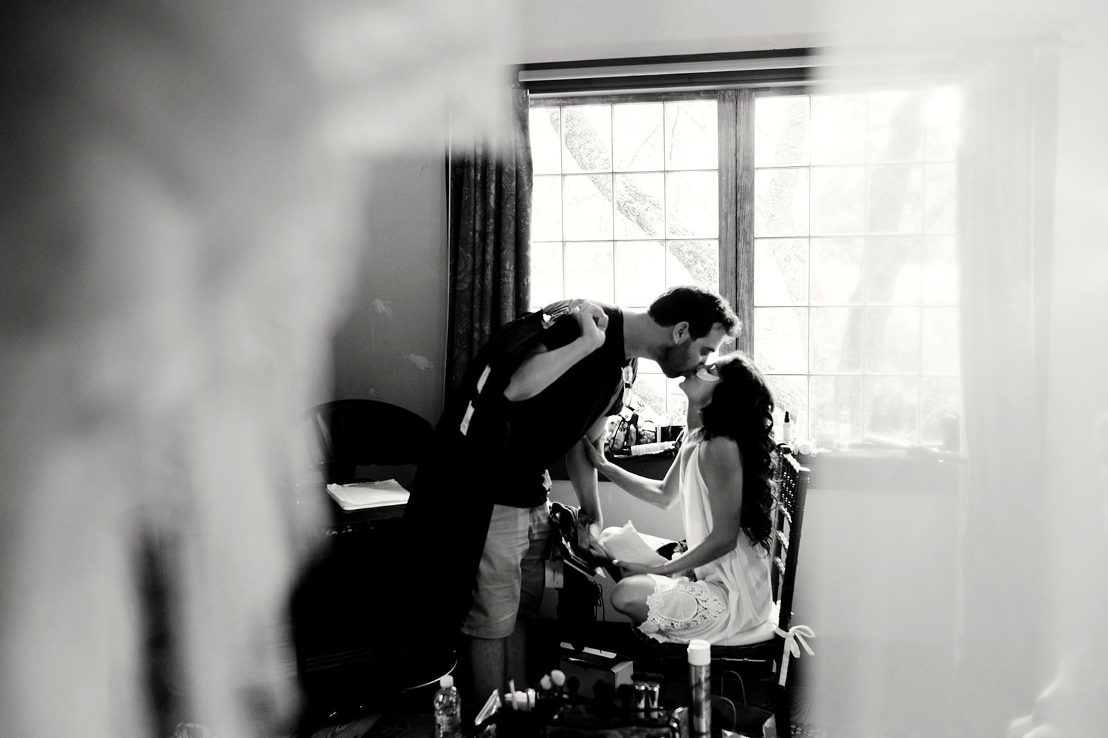A man leans over a woman sitting on a chair in front of a large window, sharing a kiss in a black and white photo.