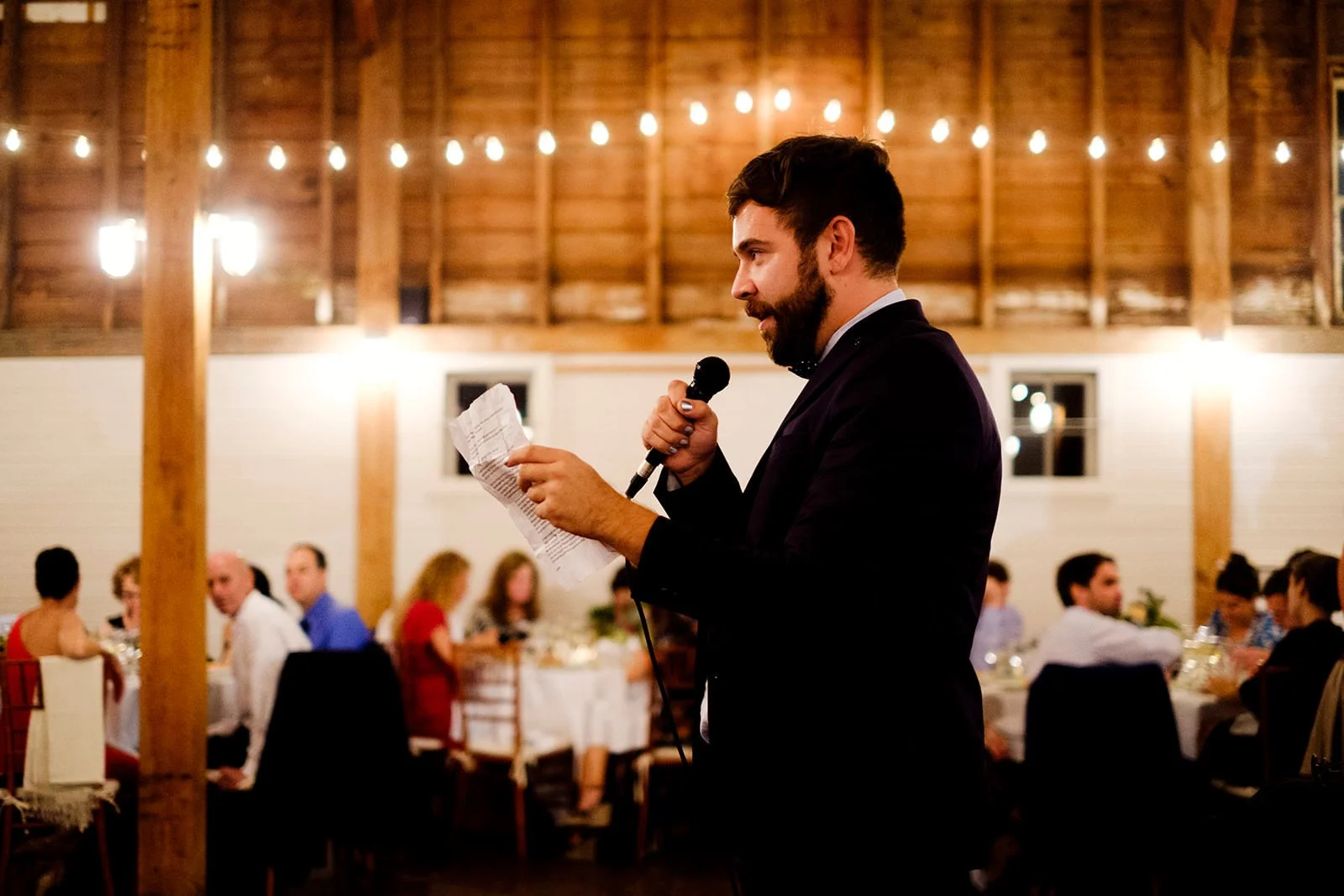 A man in a dark suit giving a speech at a wedding reception with string lights hanging from the ceiling.