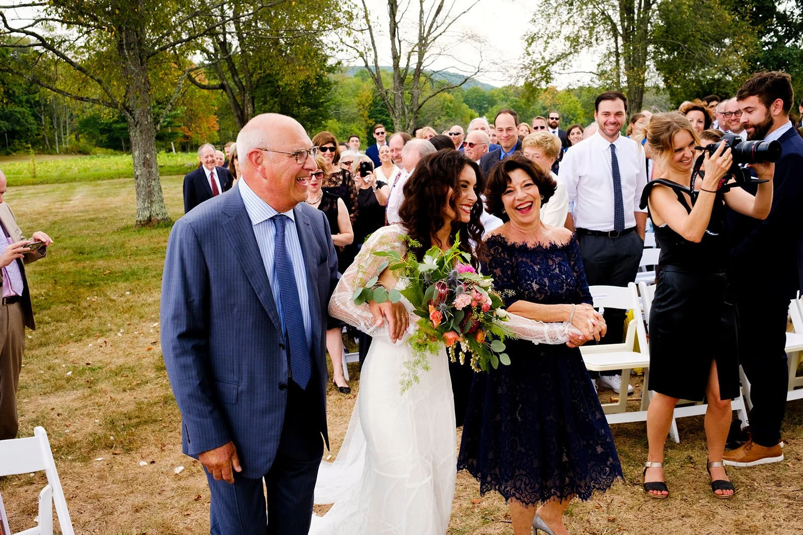 Joyful bride walking down the aisle with her parents at an outdoor wedding ceremony, surrounded by smiling guests and lush green trees.