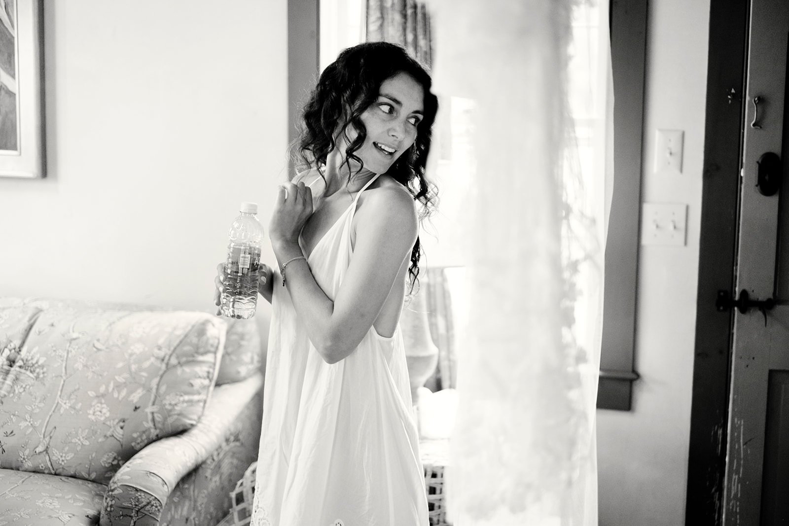 A woman in a white dress holds a water bottle and poses playfully indoors near a window with light curtains.