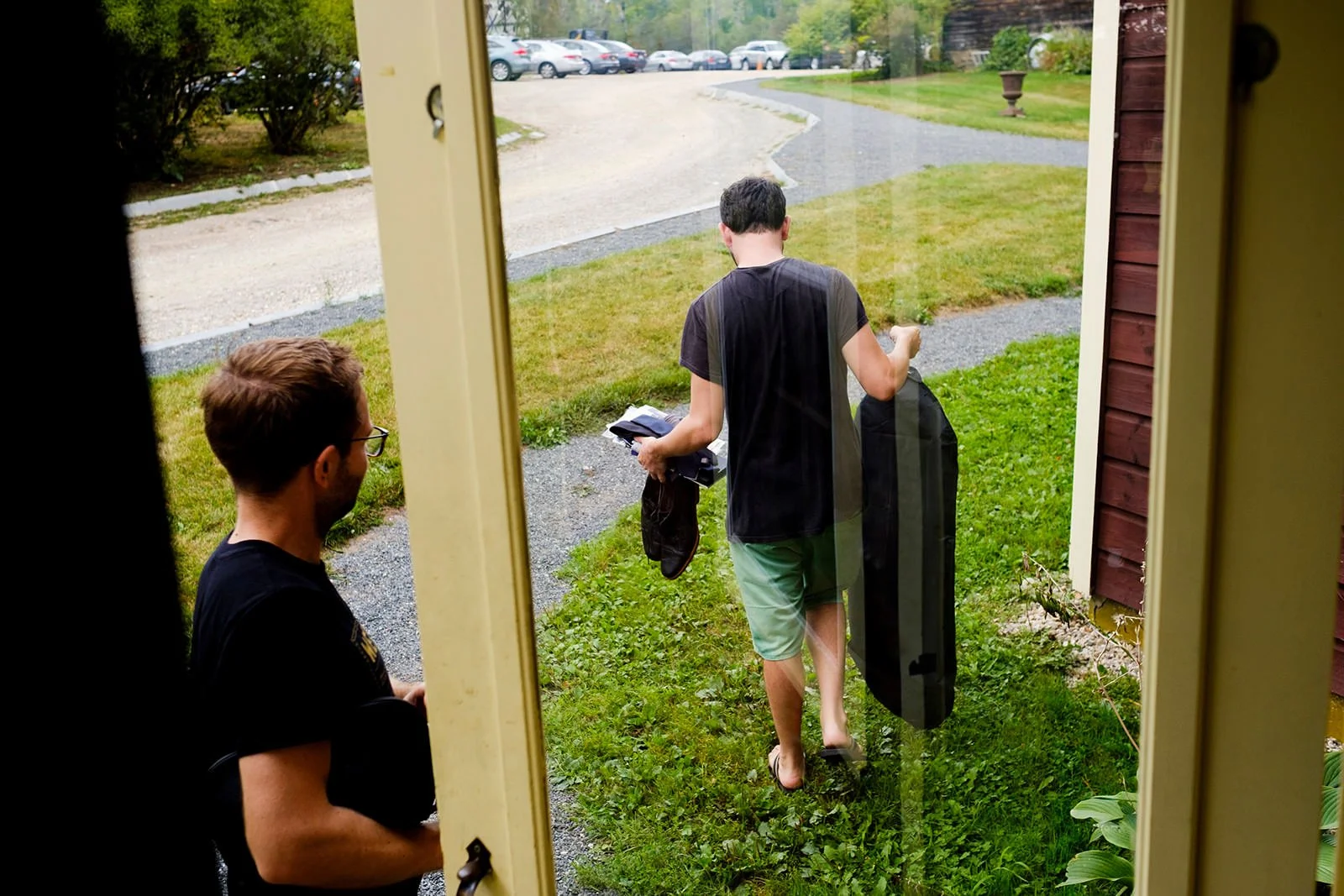 Two men are seen from a doorway, one carrying a guitar case and a bag, walking outside on a grassy area with a gravel path, towards parked cars in the distance.