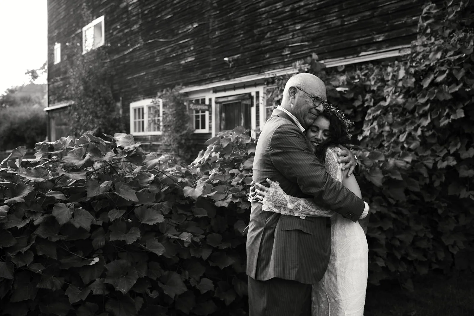 An elderly man and a young woman hugging outdoors, with a rustic house and large leafy plants in the background, in black and white.