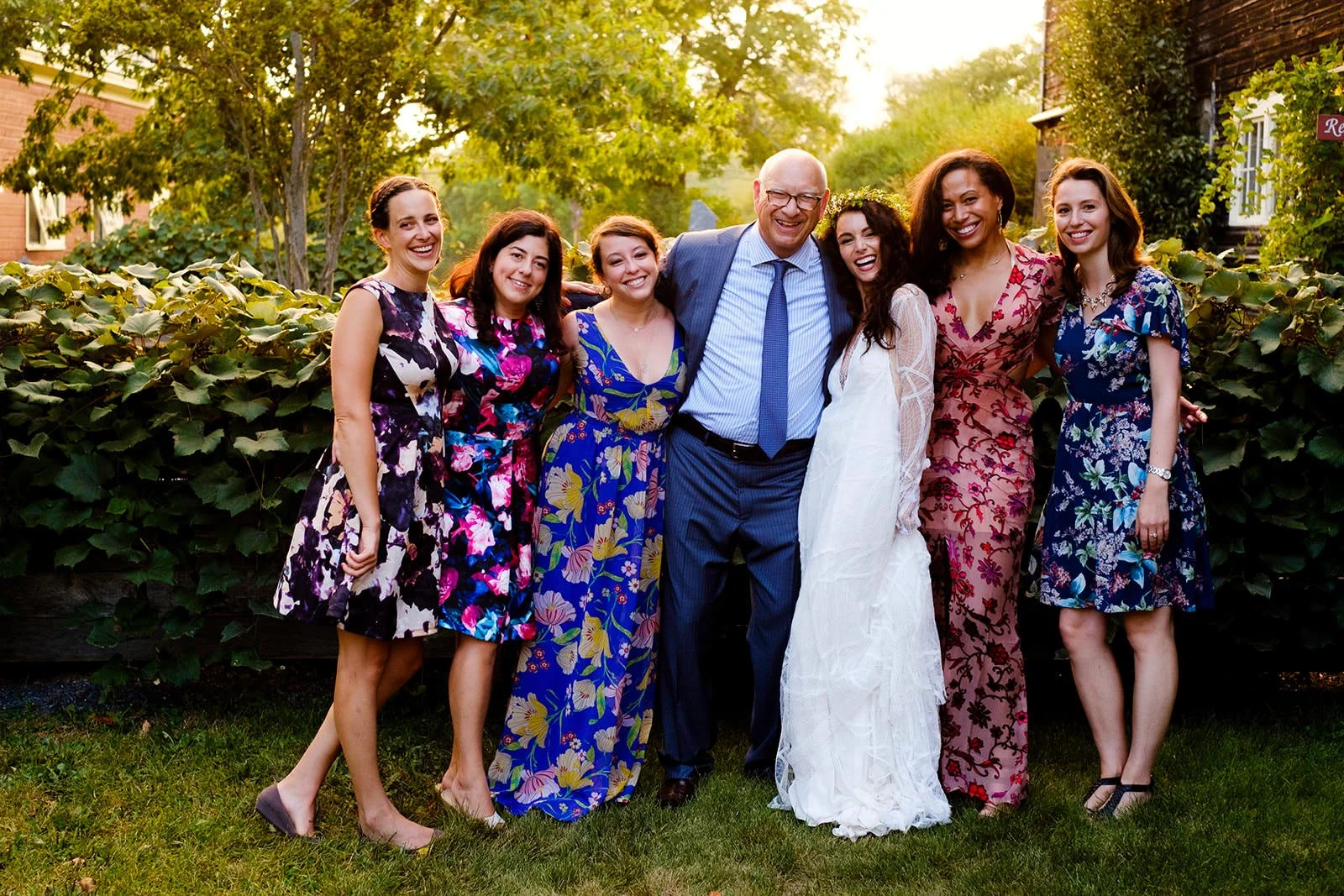 A group of seven people, six women and one man, standing together outdoors with green bushes and trees in the background during daylight. They are smiling and posing for a photo.