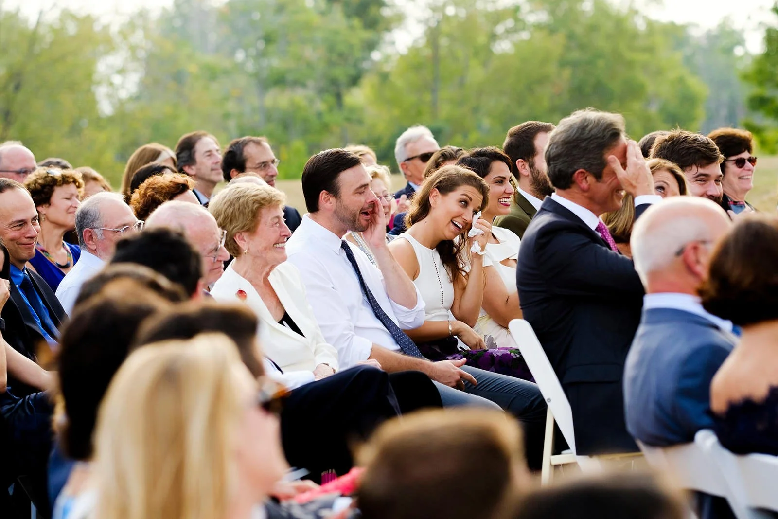 Group of people seated outdoors, attending a formal event, smiling and laughing, with trees and greenery in the background.