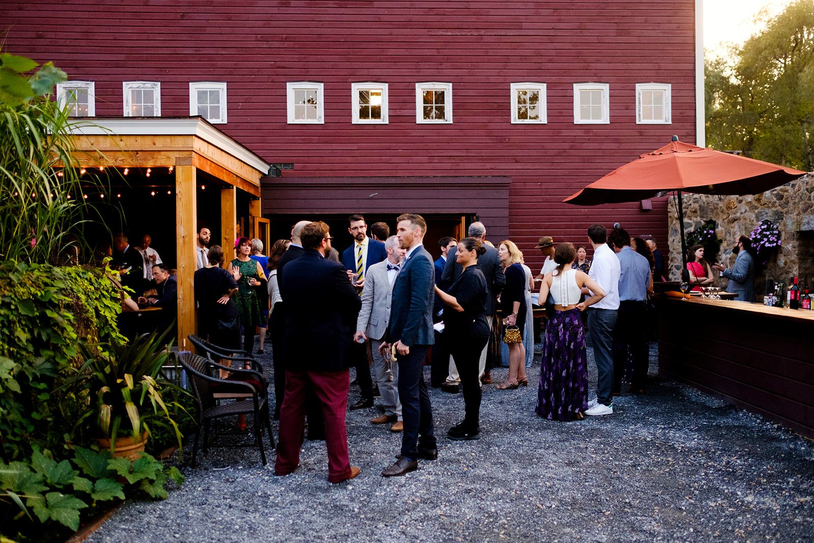 People socializing at an outdoor gathering near a red wooden building with a bar and outdoor seating under a large umbrella.