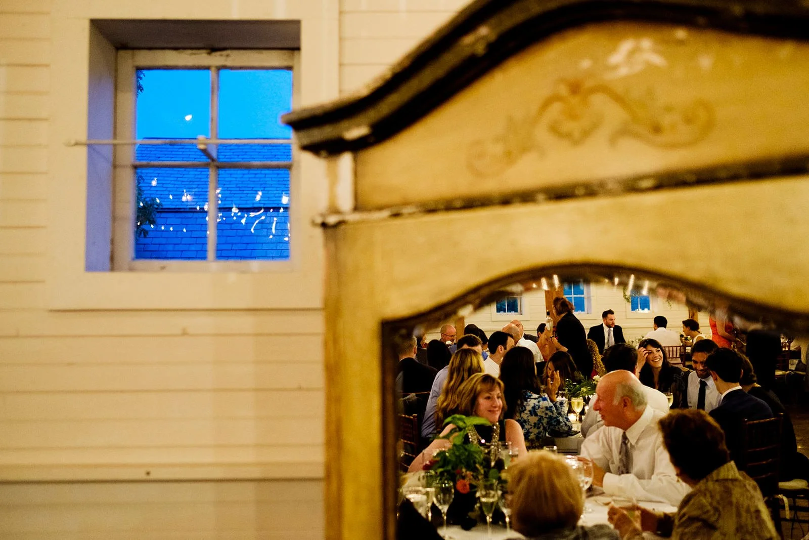 People gathered around a dinner table at an indoor event, viewed through a mirror on a wall. The room has large windows, and there are string lights outside visible through the window.