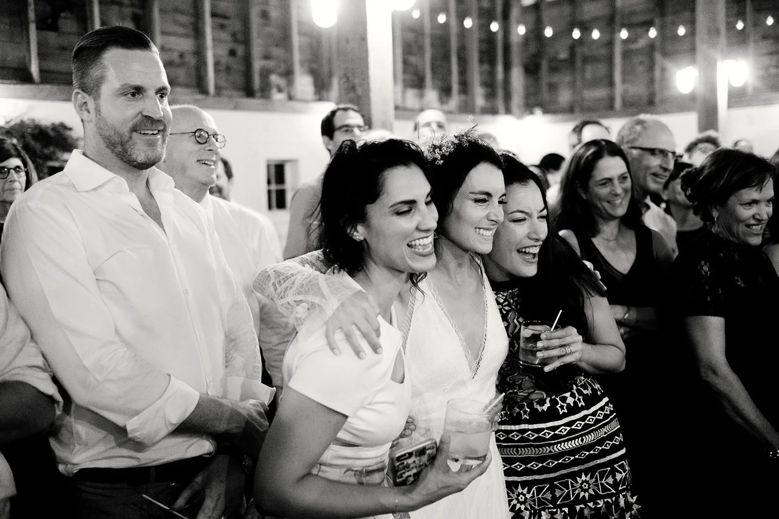 Group of people at a social gathering, smiling and laughing, in a wooden venue with string lights overhead, black-and-white photo.