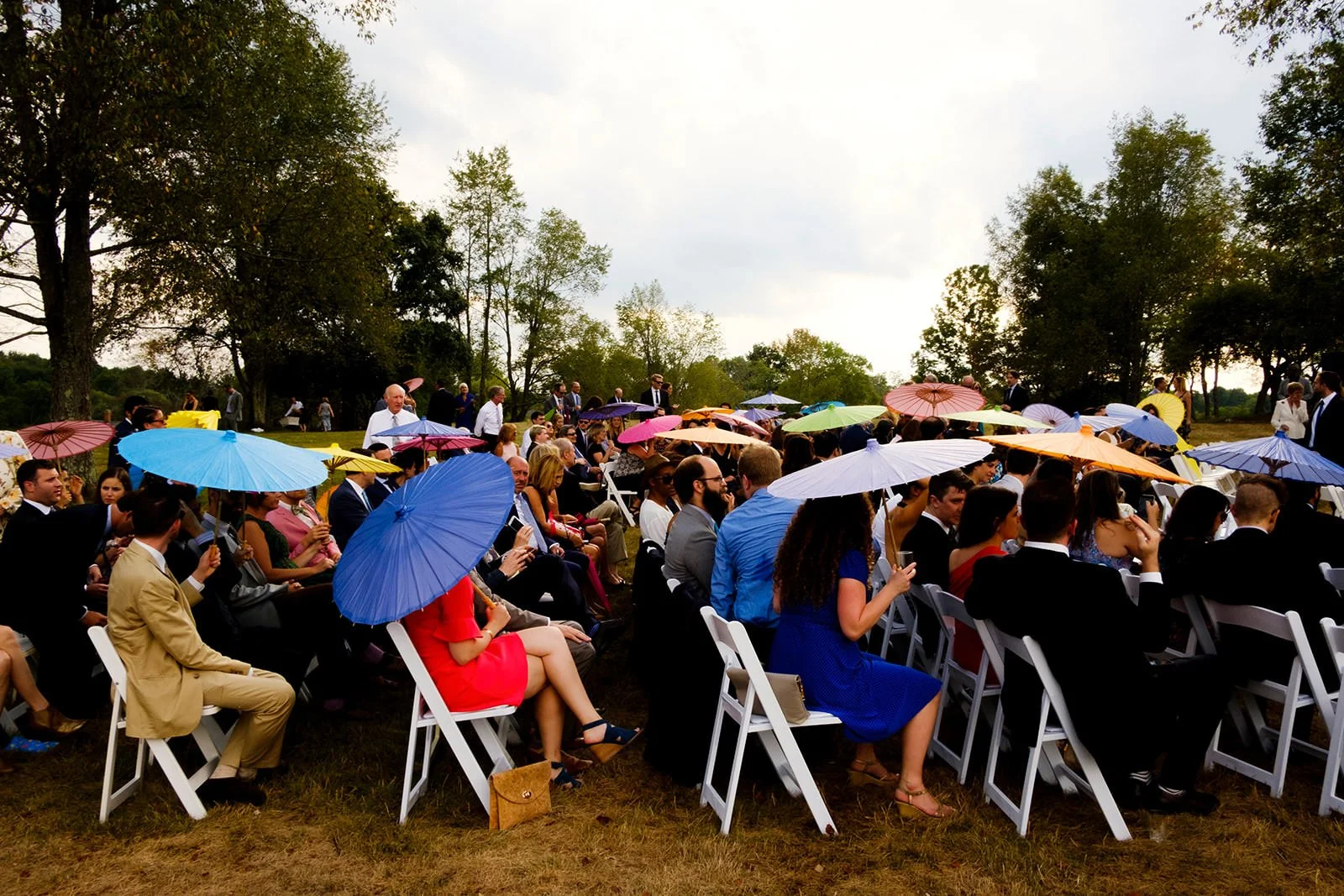 People sitting outdoors at a celebration or event, holding colorful umbrellas under a cloudy sky, with trees in the background.