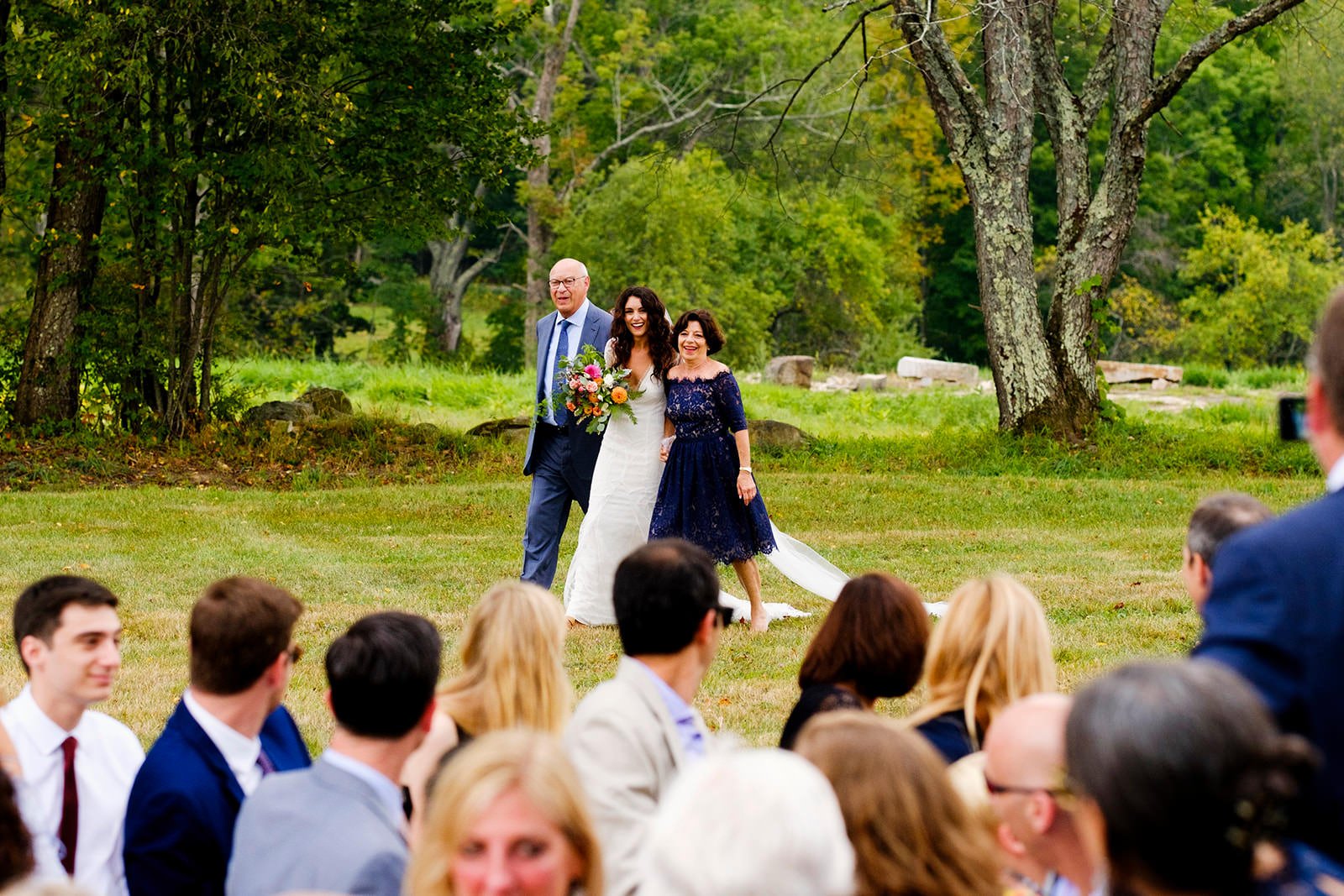 A bride in a white wedding dress holding a bouquet is walking with her parents through an outdoor wedding ceremony with guests seated on the grass under large trees.