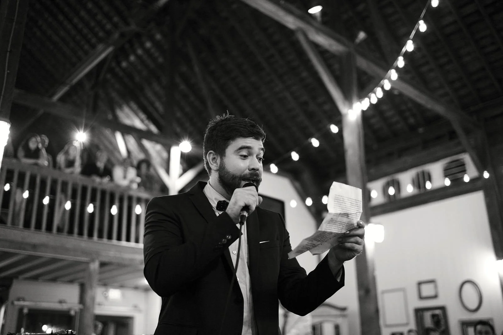 A man in a suit holding a microphone and reading from a paper, performing at an indoor event with string lights and a wooden ceiling.
