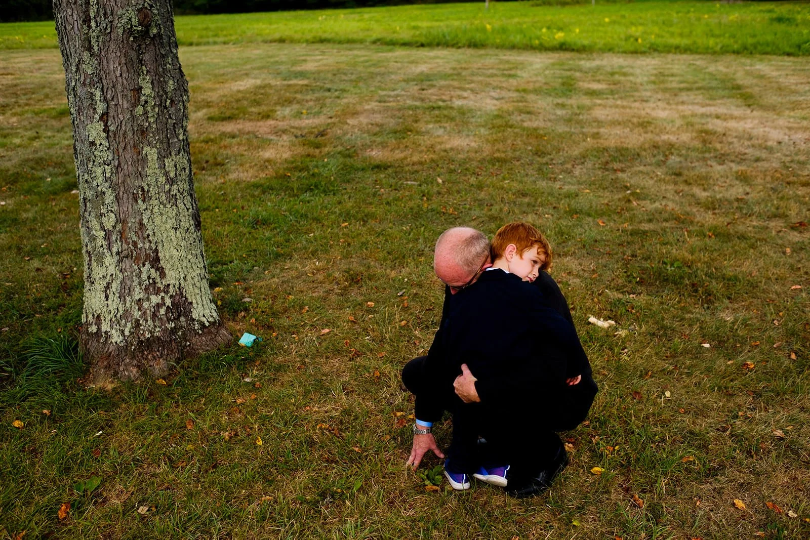 A man and a young boy hugging on a grassy area near a tree in a park.