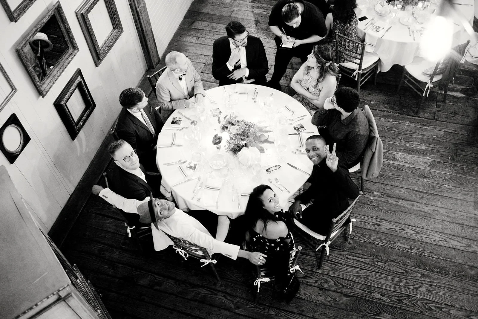 A group of people sitting around a round banquet table at a formal event, smiling and posing for the camera, with some making peace signs, in a wooden-floored room decorated with framed pictures on the wall.