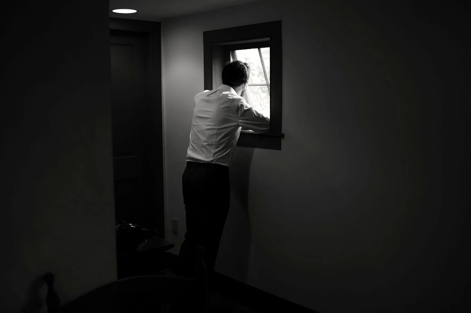 A man in a white shirt stands by a small window, looking outside, in a dimly lit room.