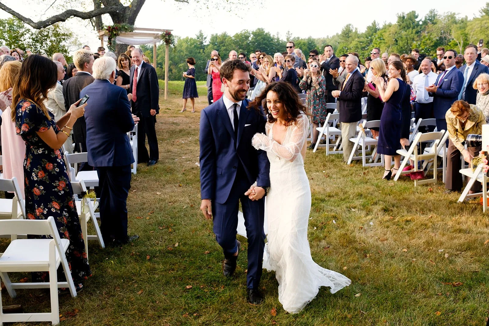 A bride and groom walking hand in hand down the aisle after their wedding ceremony, surrounded by seated and standing guests outdoors on a grassy field.